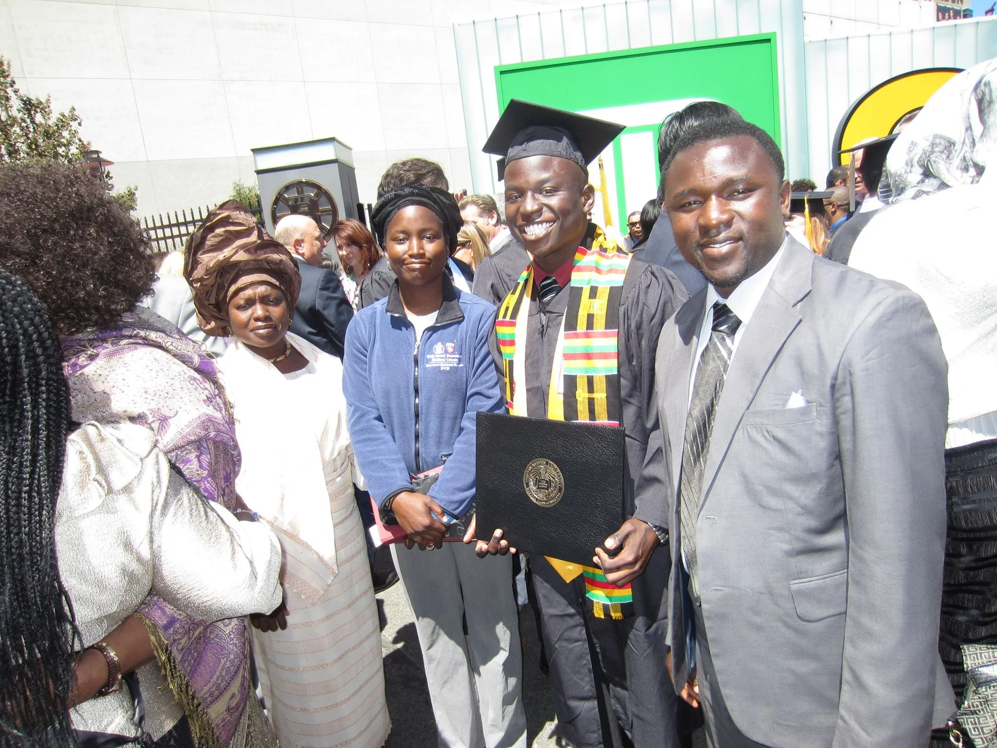 A diverse group of people at a graduation ceremony, with one graduate in cap and gown holding a diploma, surrounded by family and friends outdoors.