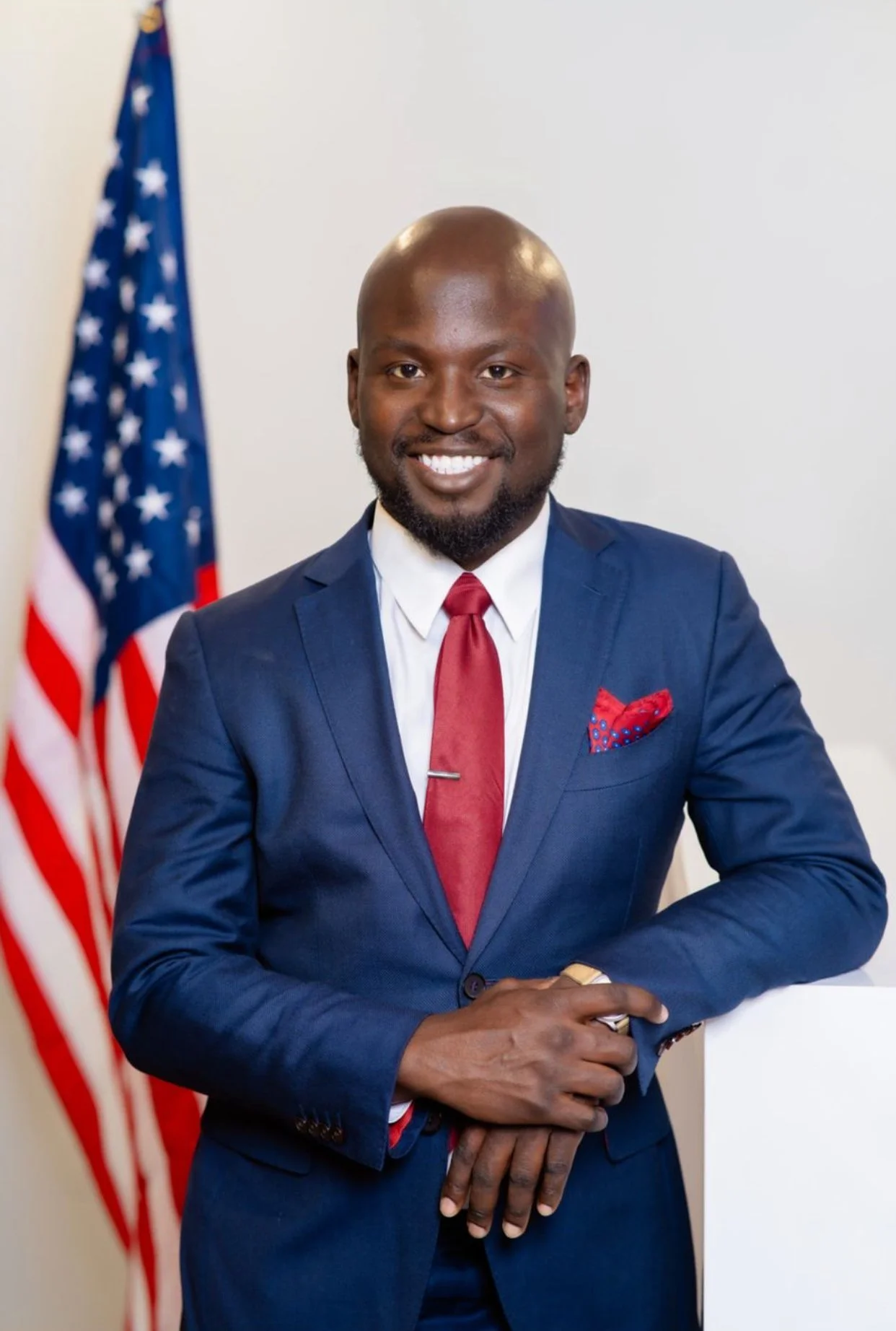 A smiling man in a blue suit, white shirt, and red tie, standing next to an American flag.