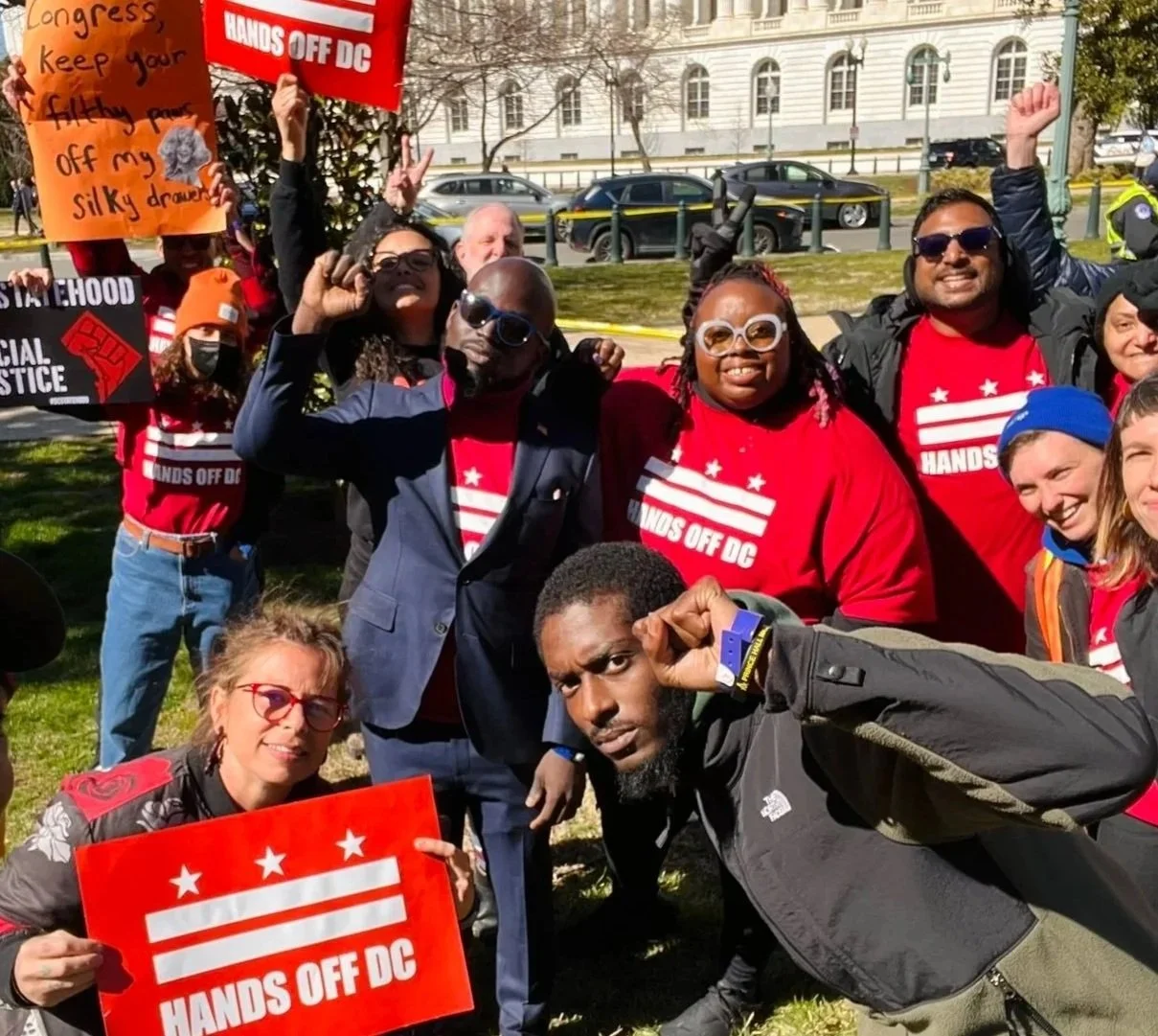 Group of protesters outdoors, holding signs that read "HANDS OFF DC" and "Congress keep your filthy paws off my silky drawers". People are smiling, making peace signs, and wearing red shirts with white stripes and stars.