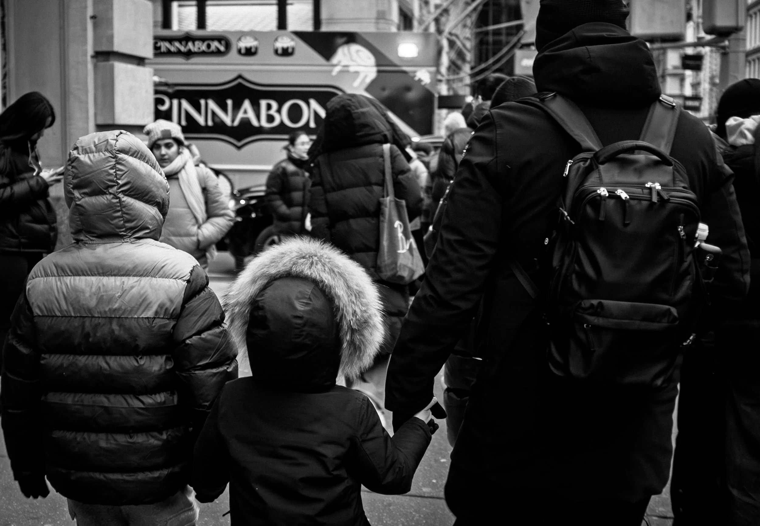 Black and white photo of a crowded city street with people dressed in winter clothing, including jackets and hoodies. Some are wearing backpacks. A sign for 'PINNABON' is visible in the background.