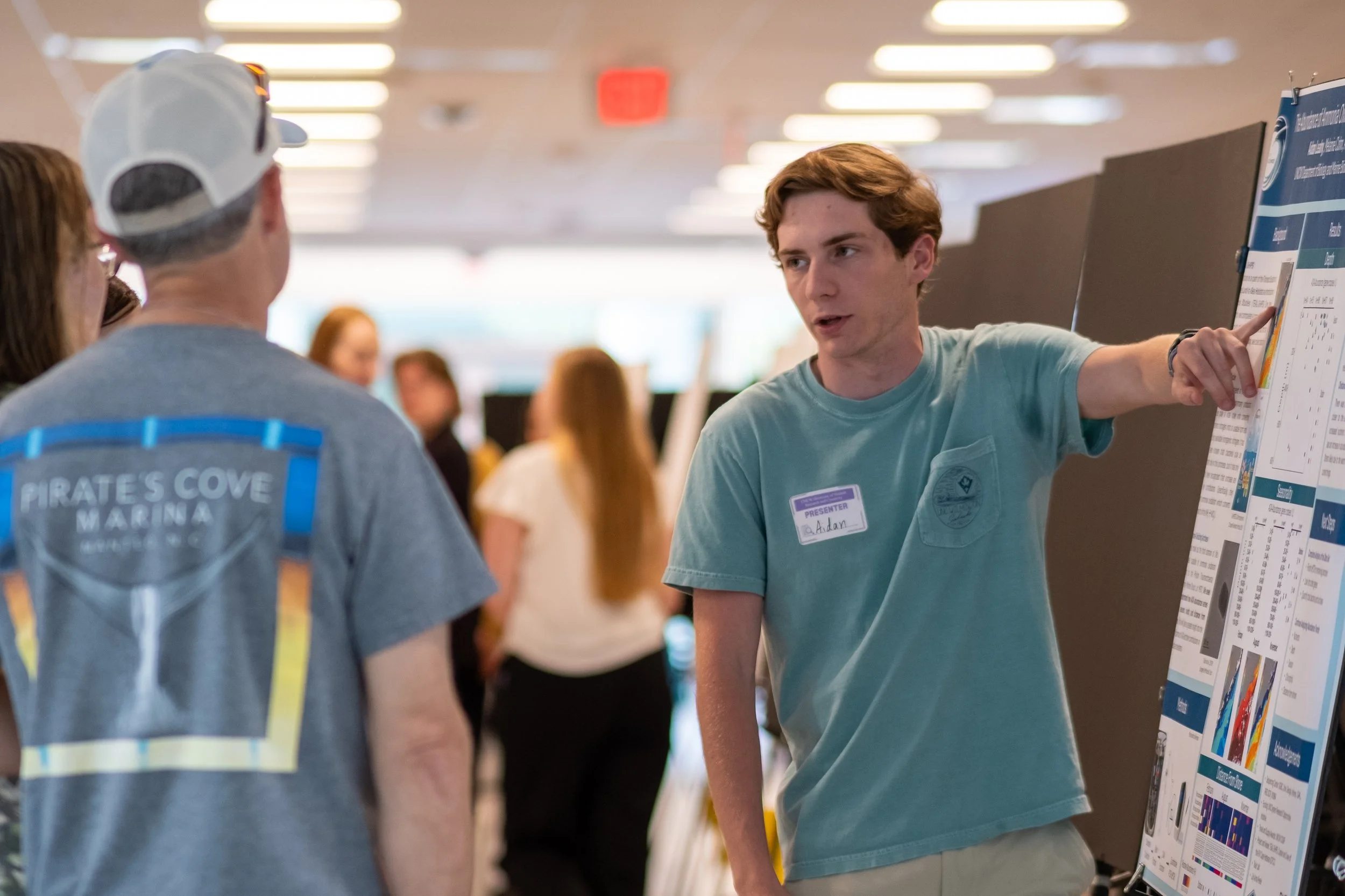 A young man with a name tag labeled 'Presenter' is speaking to a group of people at an indoor event. The young man is pointing to a large poster or display board with charts and information. The background shows other attendees and a brightly lit roo