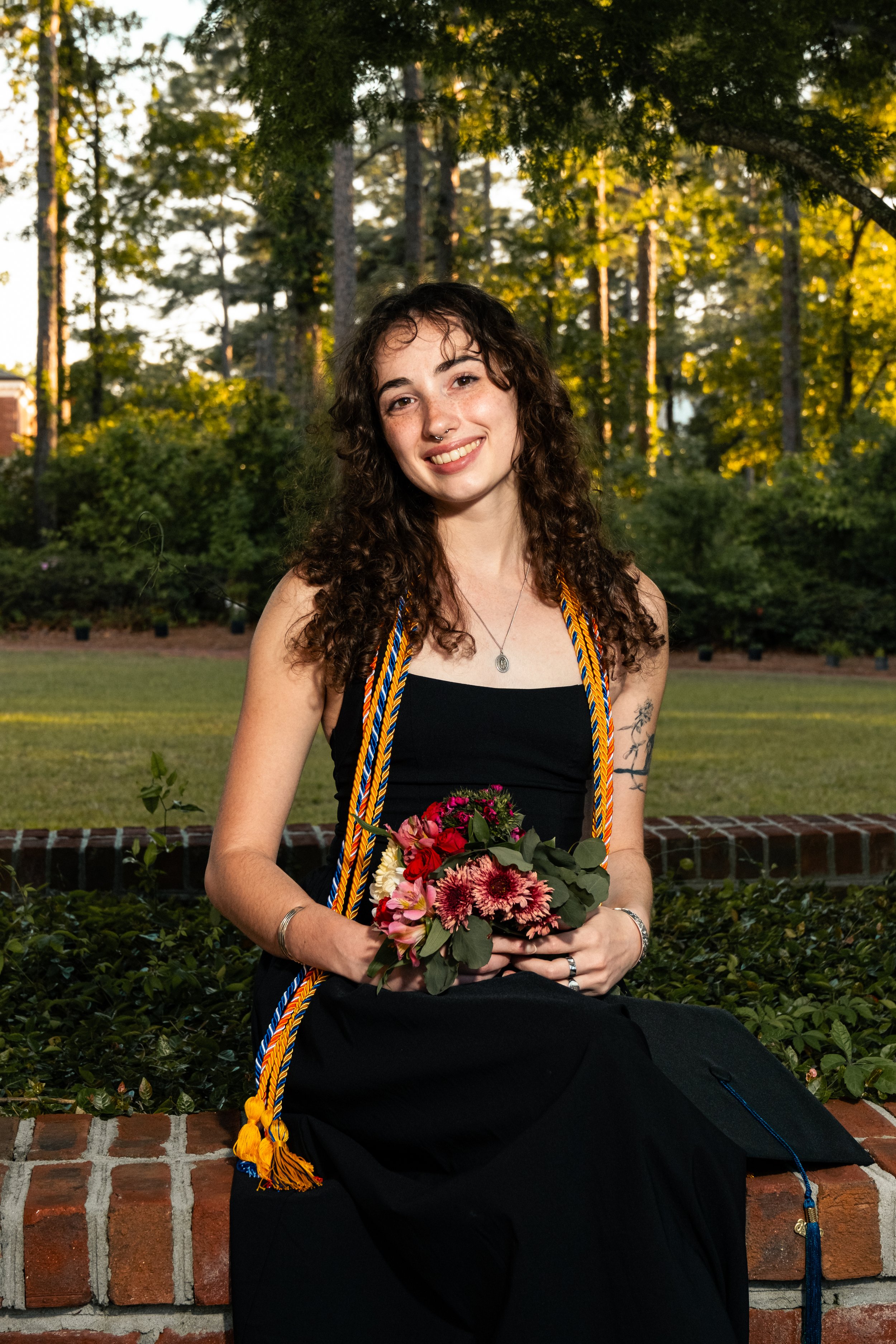 Young woman with curly hair and a nose piercing holding a bouquet of flowers, wearing a black dress and graduation cords, sitting outside on a brick wall during sunset with a background of trees and greenery.