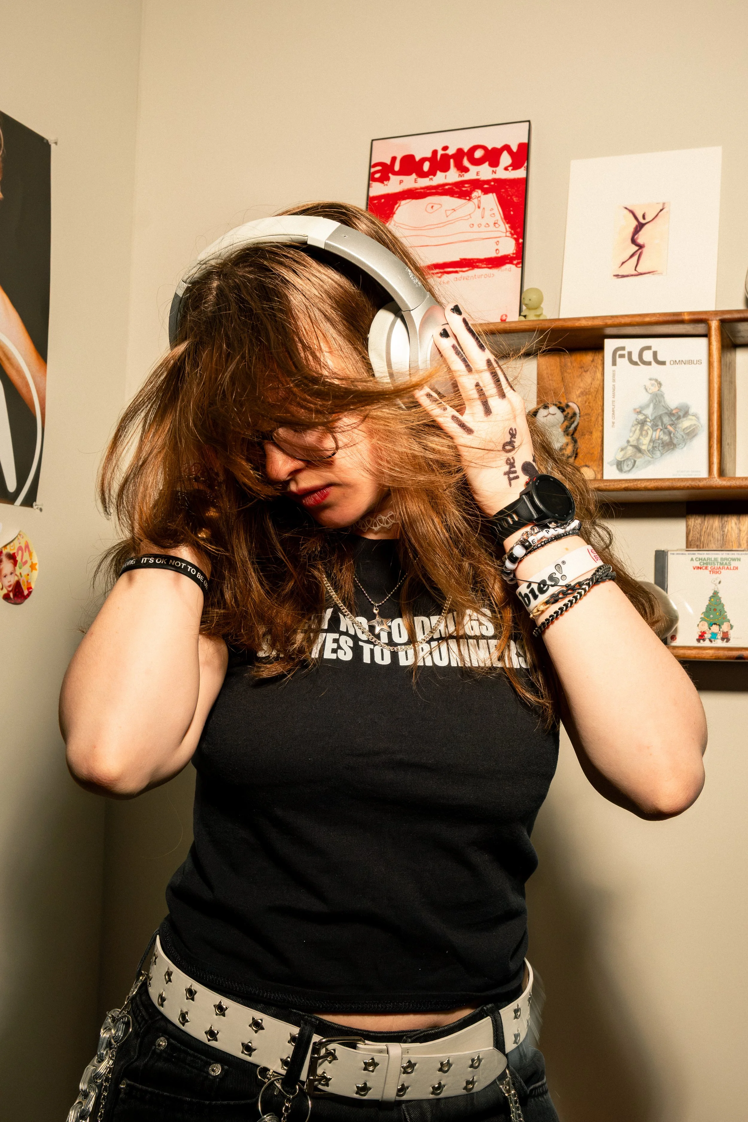 A person with brown hair wearing headphones, a black T-shirt, and a white belt with studs, standing in front of a beige wall with various posters and artwork.