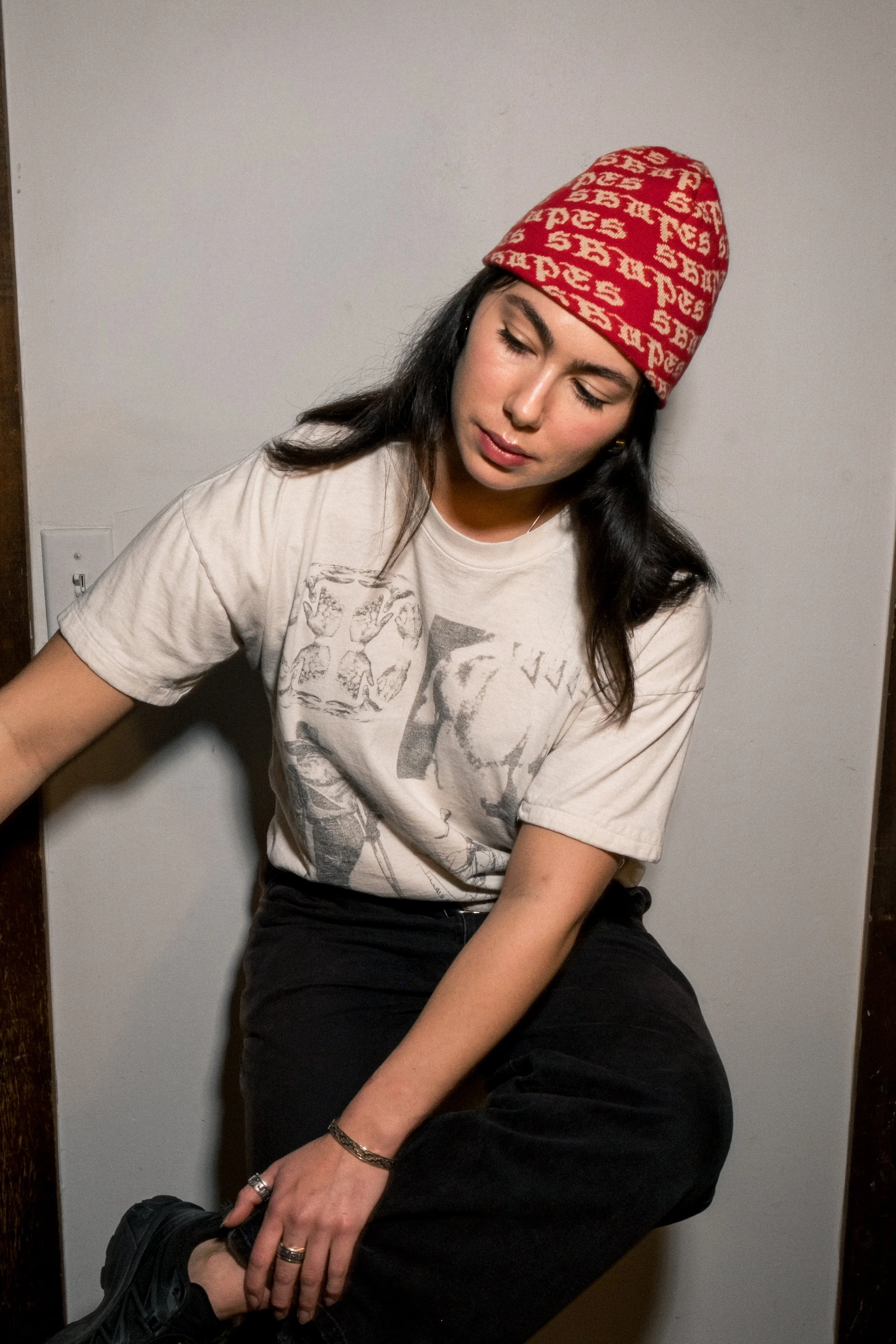 A young woman with long dark hair, wearing a red beanie with white lettering, a graphic t-shirt, and black pants, sitting against a plain wall, looking down and holding her ankle, with a neutral expression.