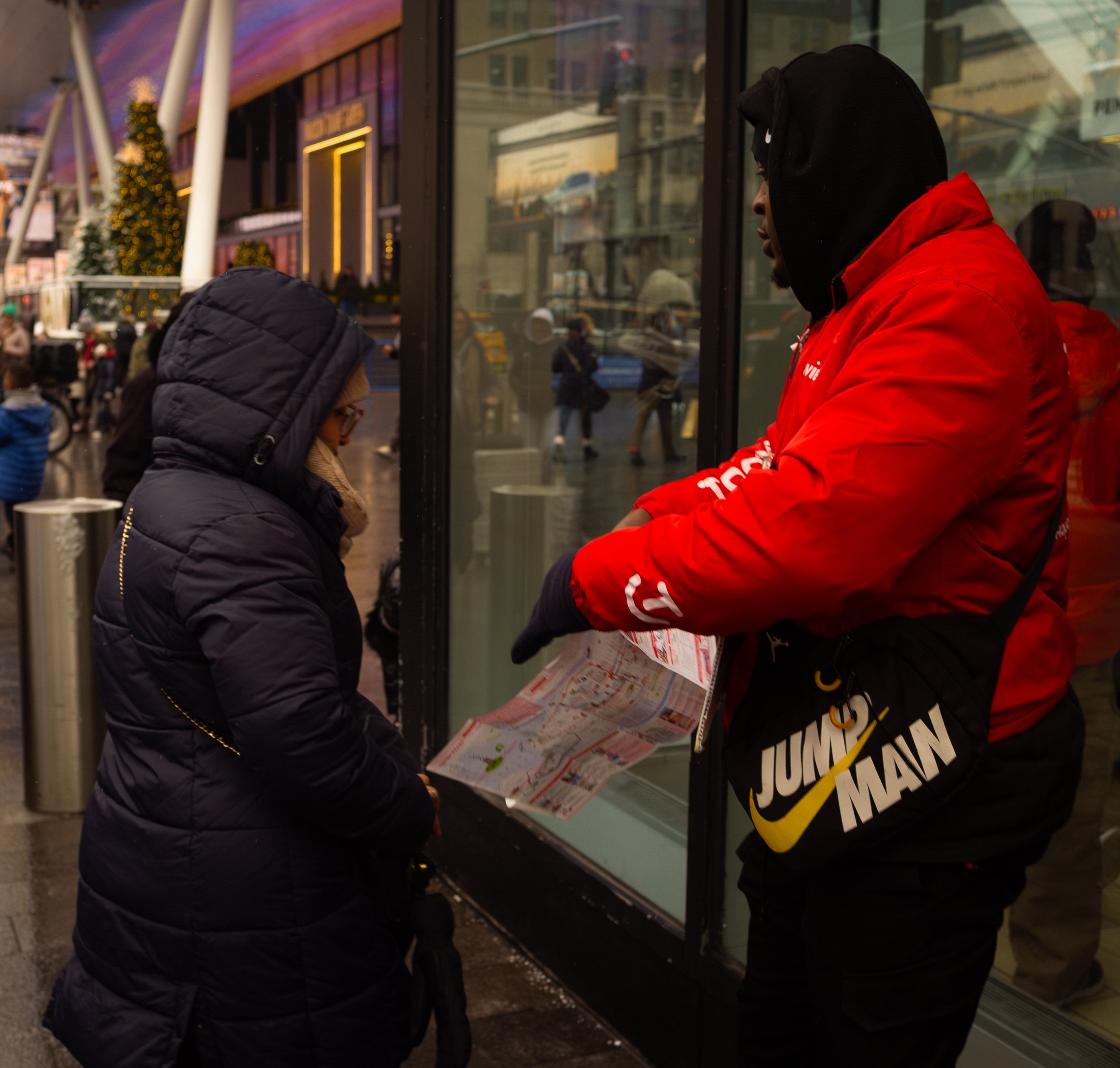 A person dressed in a black winter coat with a hood, glasses, and beige scarf talking to a street vendor through a glass window. The vendor is wearing a red jacket and black gloves, holding a map or brochure, with a black Nike Jumpman bag.
