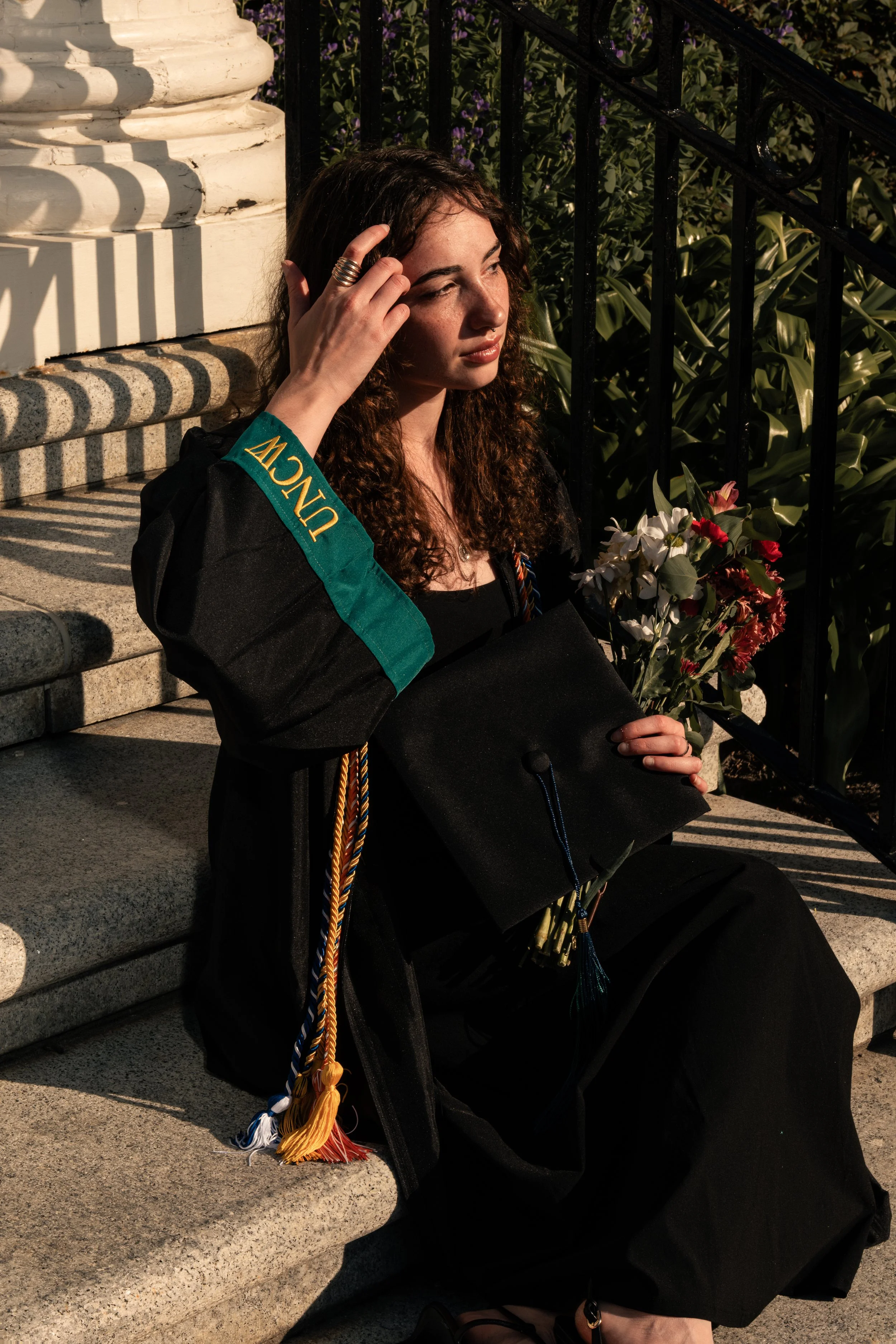 A young woman in graduation attire sitting on steps outside, holding a diploma and flowers, wearing a black graduation gown with a green collar and gold tassels