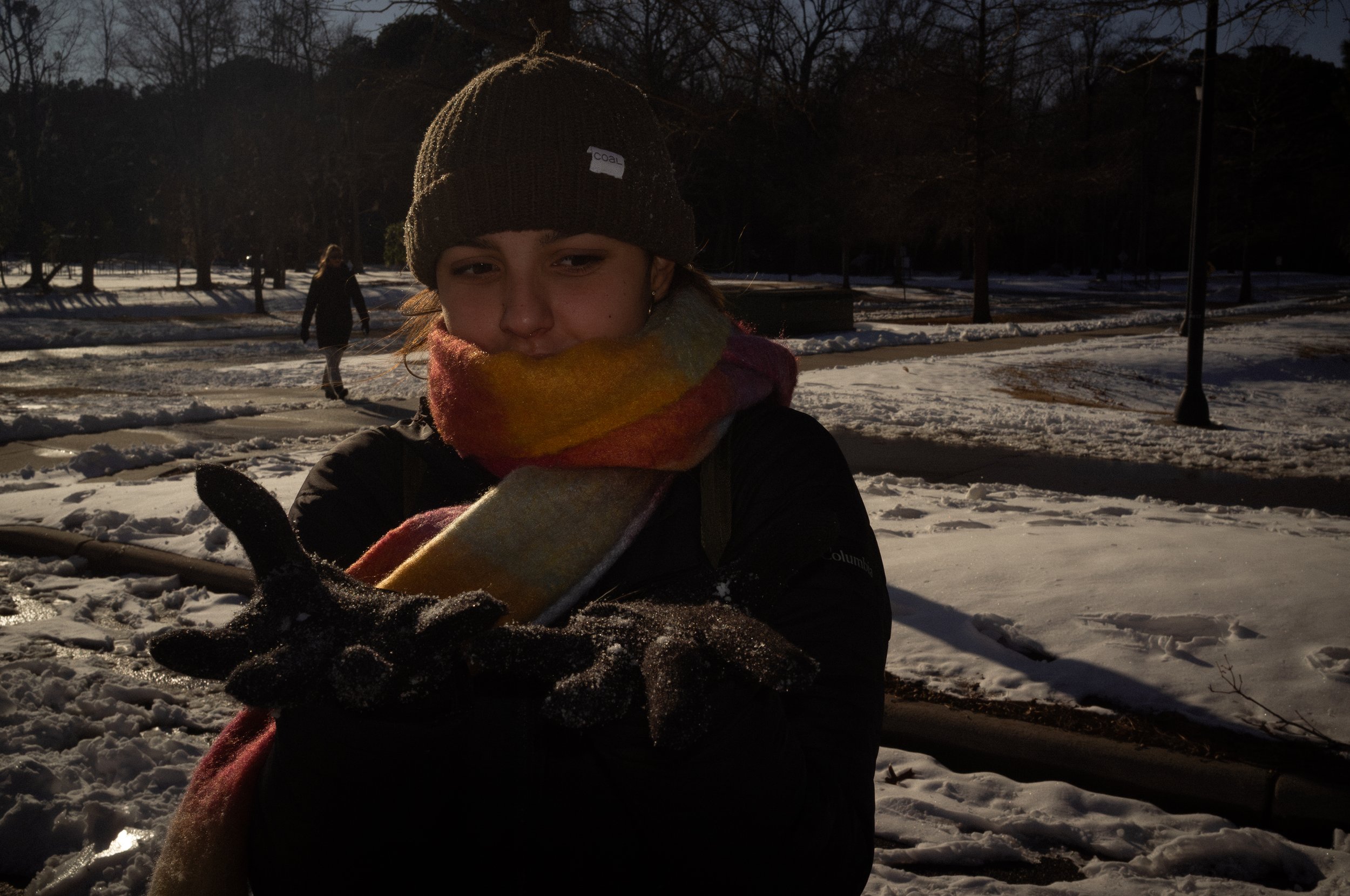 A young person with a beanie, scarf, and winter coat looking at the camera in a snowy park during daylight, with other people in the background.