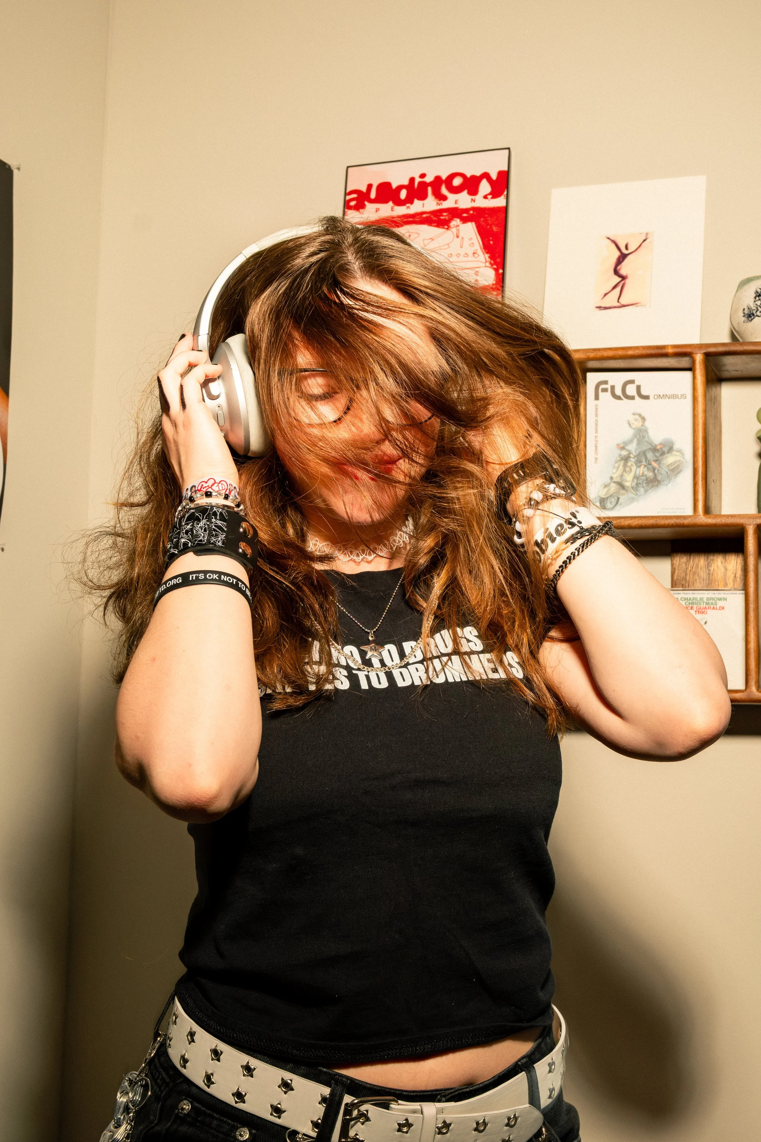 A young woman with long brown hair wearing glasses, headphones, multiple bracelets, and a black t-shirt with white text. She has a white belt with star-shaped studs and black pants with chains. She is in an indoor setting, touching her headphones, wi
