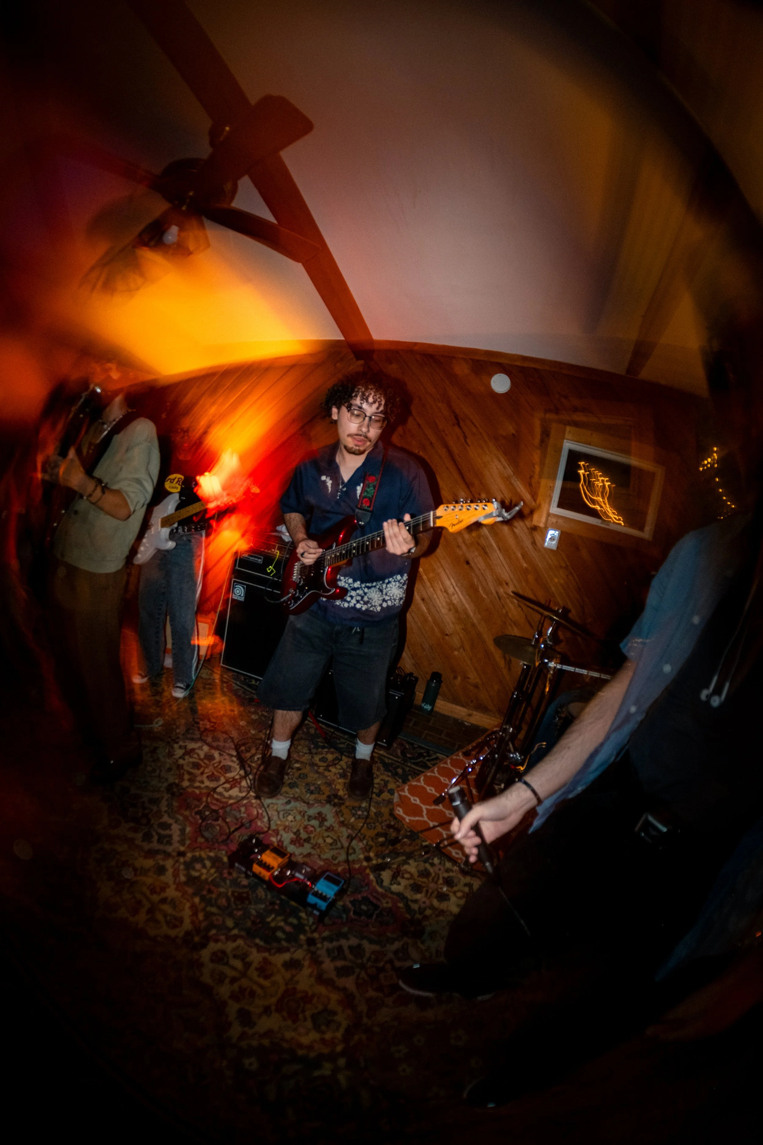 A group of young musicians playing electric guitars and a drum set in a cozy, wood-paneled room with a patterned carpet and warm lighting, captured with a fisheye lens.