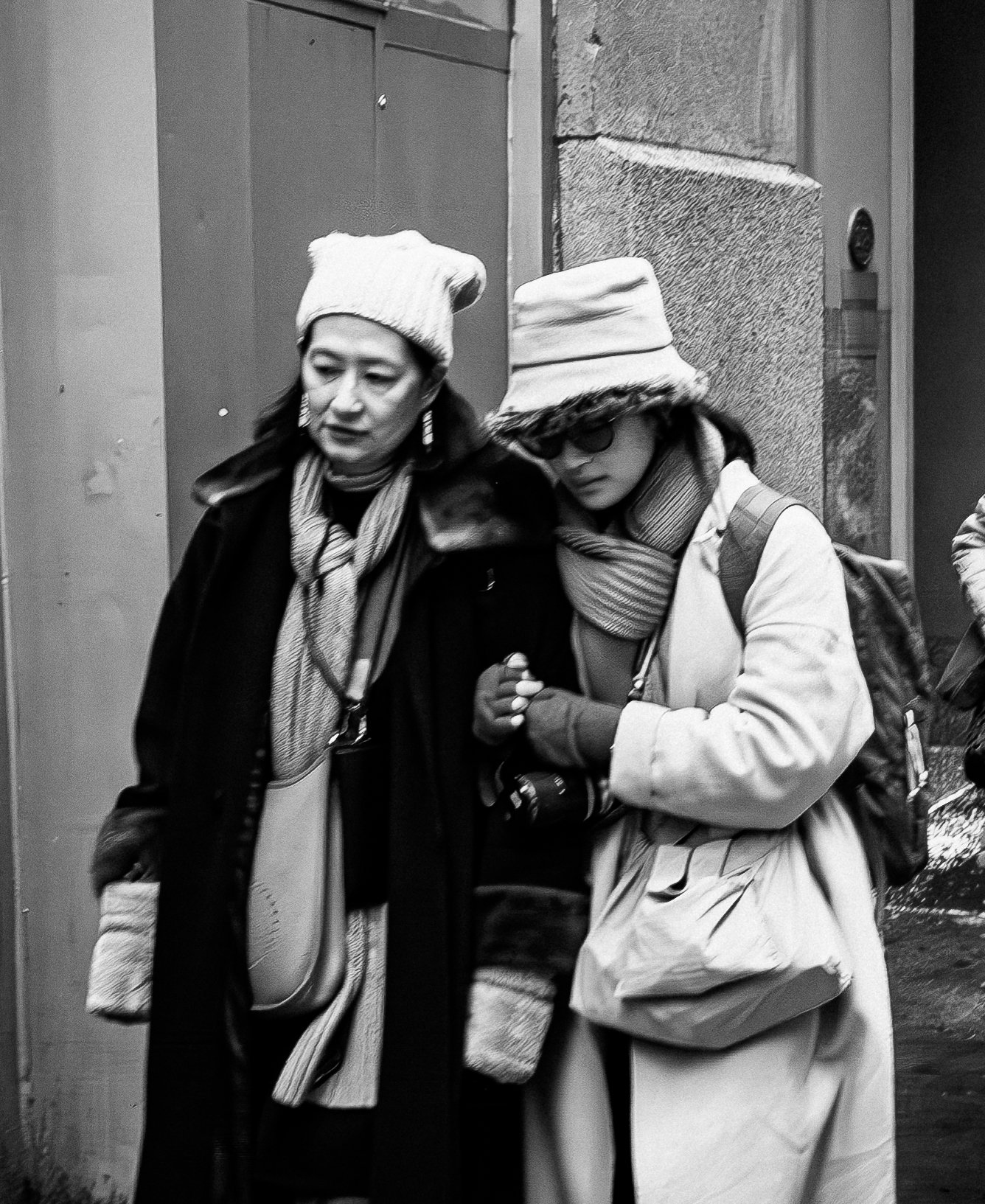 Two women wearing winter clothing and hats walk closely together on a city street at night.