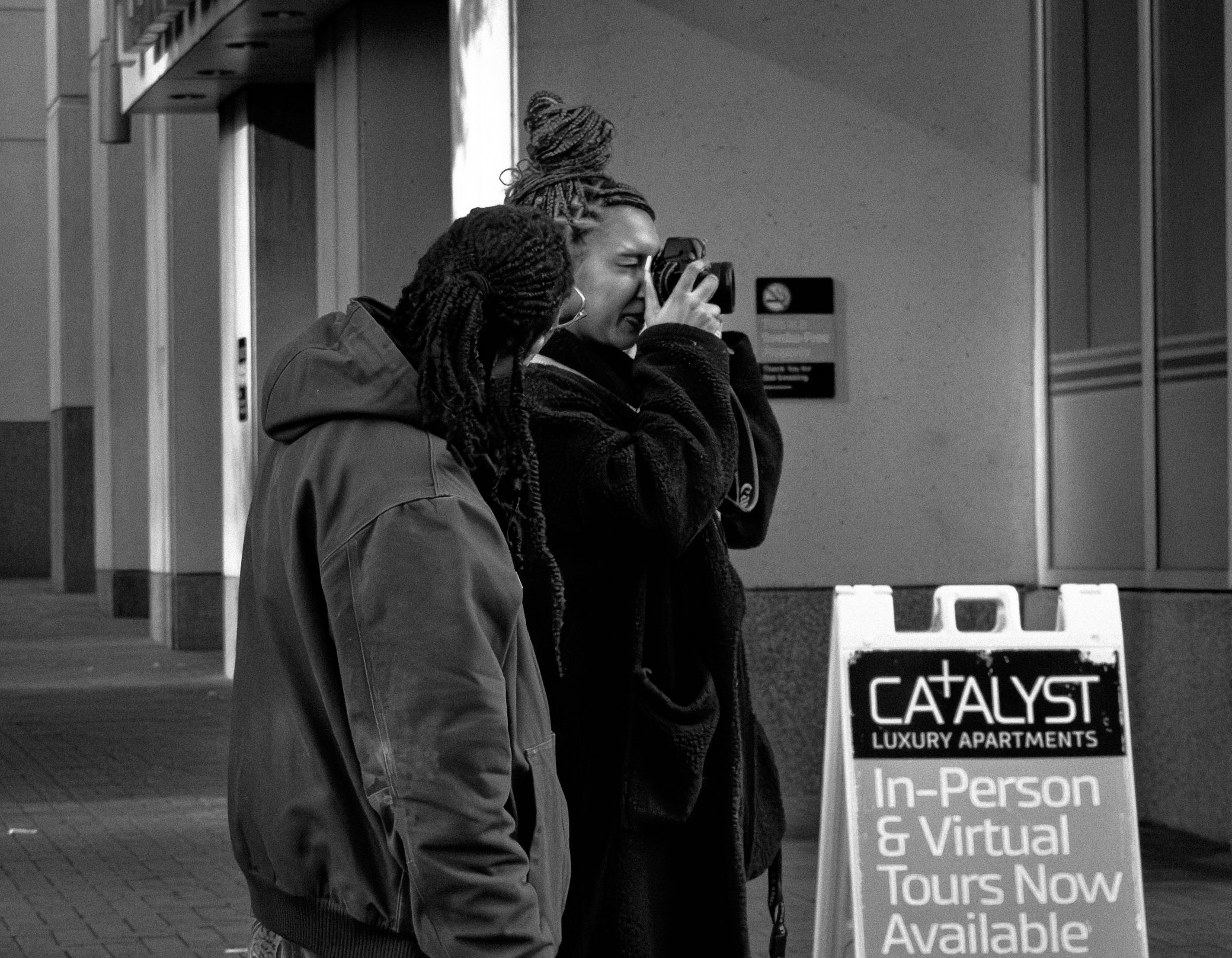 Two women with braided hair, one taking a photo with a camera, standing on a city sidewalk near an advertisement sign for Catalyst Luxury Apartments offering in-person and virtual tours.