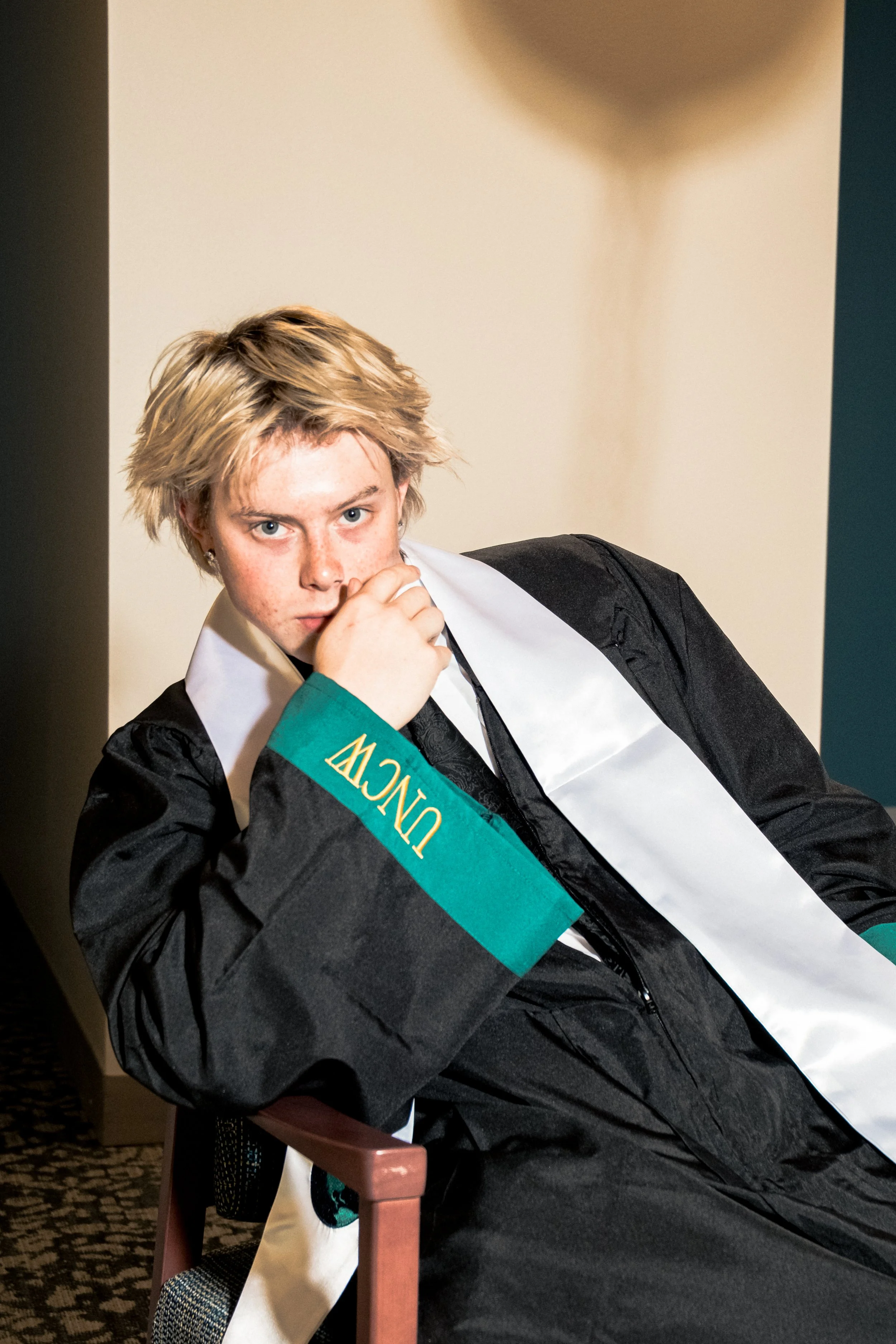 Young person with short, light-colored hair and freckles wearing a black graduation gown and white sash, sitting indoors with a beige wall behind them, looking at the camera with a serious expression.