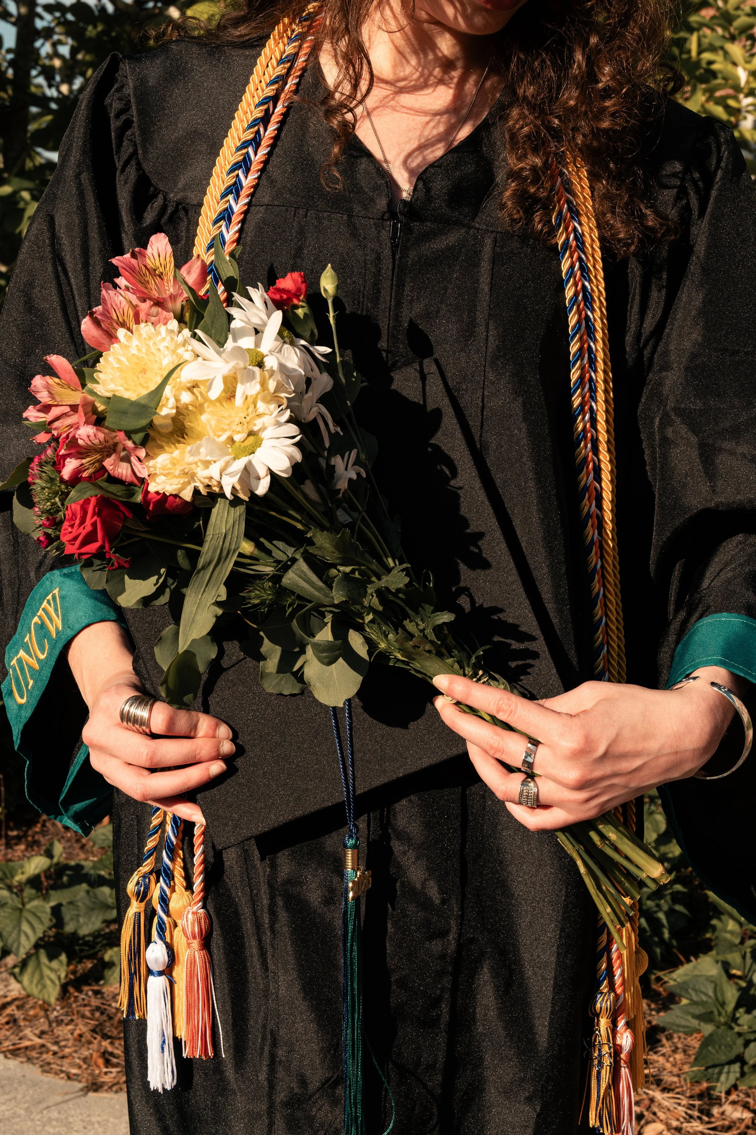 Close-up of a person wearing a black graduation gown, holding a bouquet of flowers, with honor cords around their neck, and various rings on their fingers.