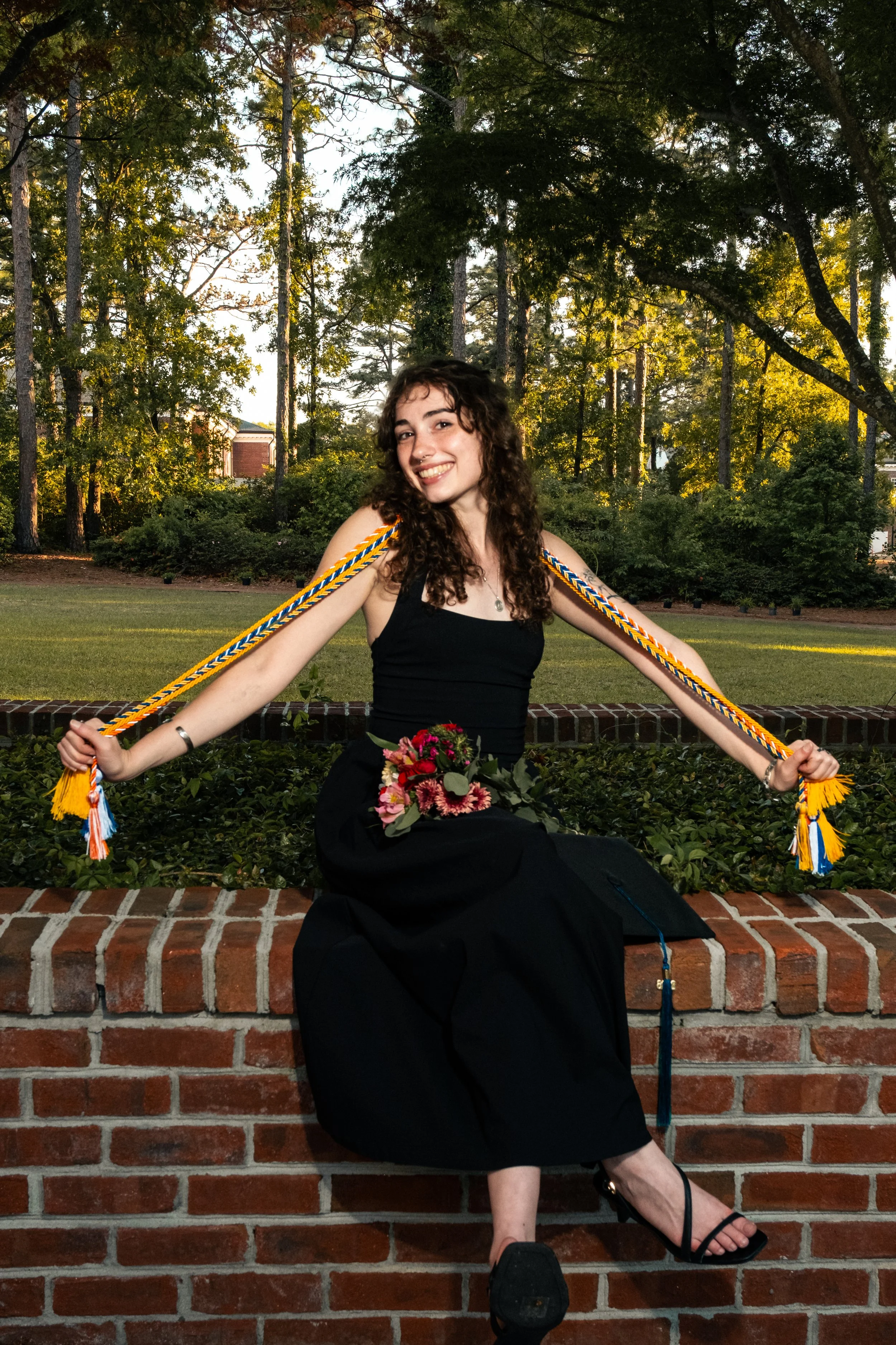 A young woman with curly hair and a black dress is sitting on a brick wall in a park, smiling and holding colorful cords with tassels over her shoulders. There are trees and a building in the background during sunset.