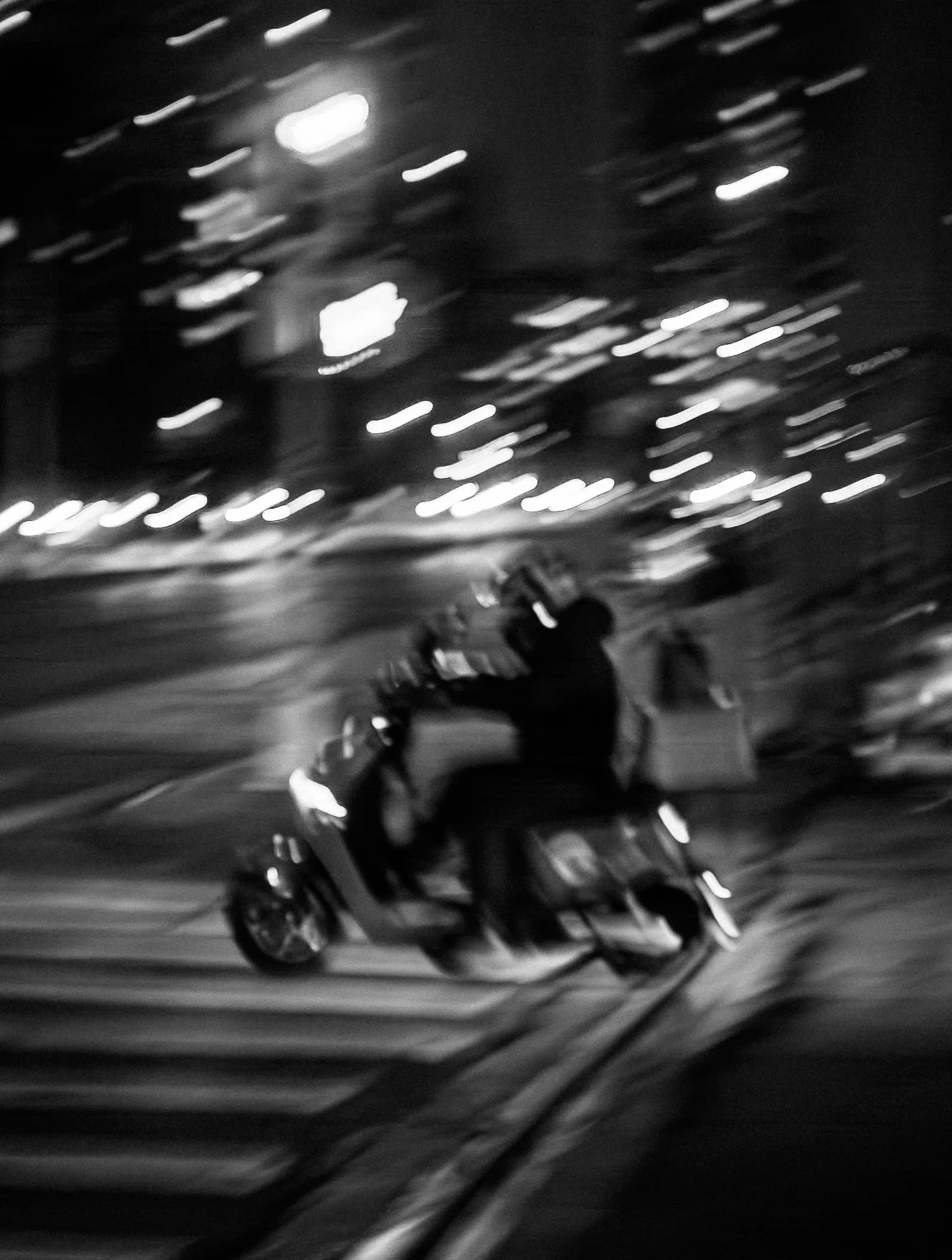 Black and white photo of a person riding a scooter on a city street at night, with blurred lights and motion in the background.