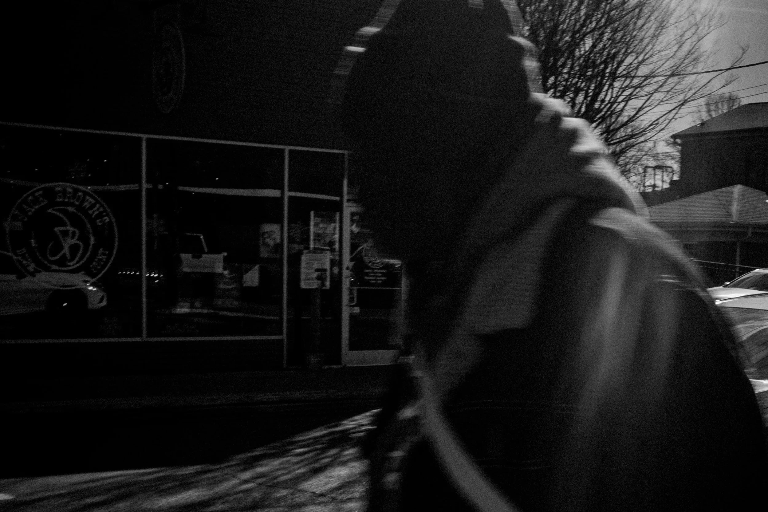 A person walking outside at night near a glass storefront with a logo that says 'The Bright Side'. The person's face is not visible, and there are parked cars and a house with trees in the background.
