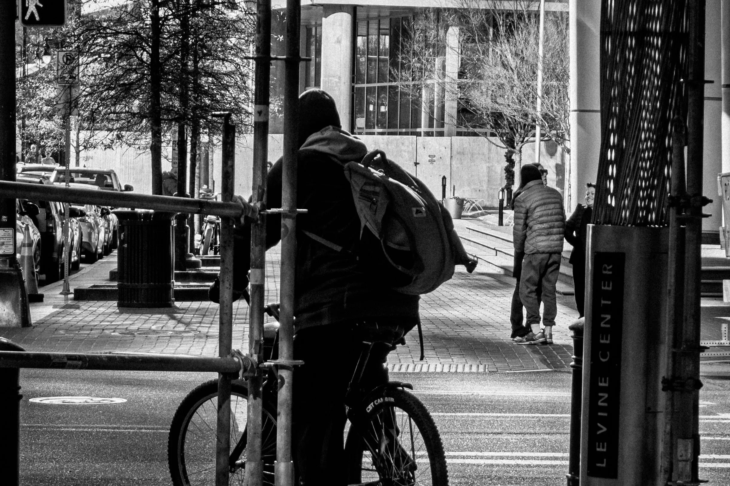 A person with a backpack on a bicycle waiting behind a construction barricade on an urban street, with two other people walking in the background near a sidewalk and modern buildings.