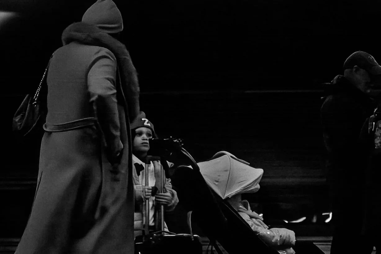 A woman standing next to a young boy in a stroller on a dark street at night, with another person walking nearby.