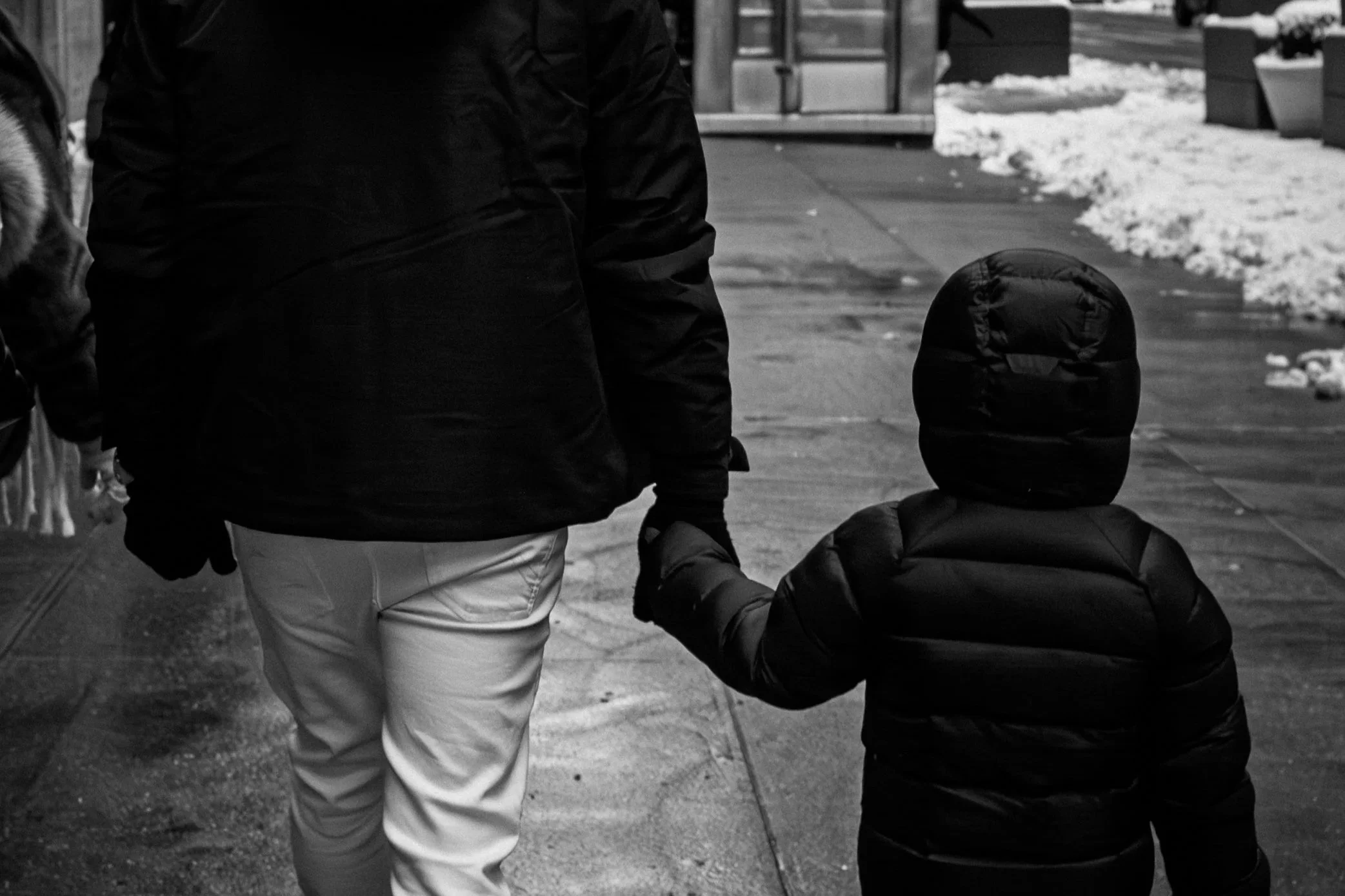 A person holding a child's hand while walking on a city sidewalk in winter, with snow on the ground and storefronts in the background.