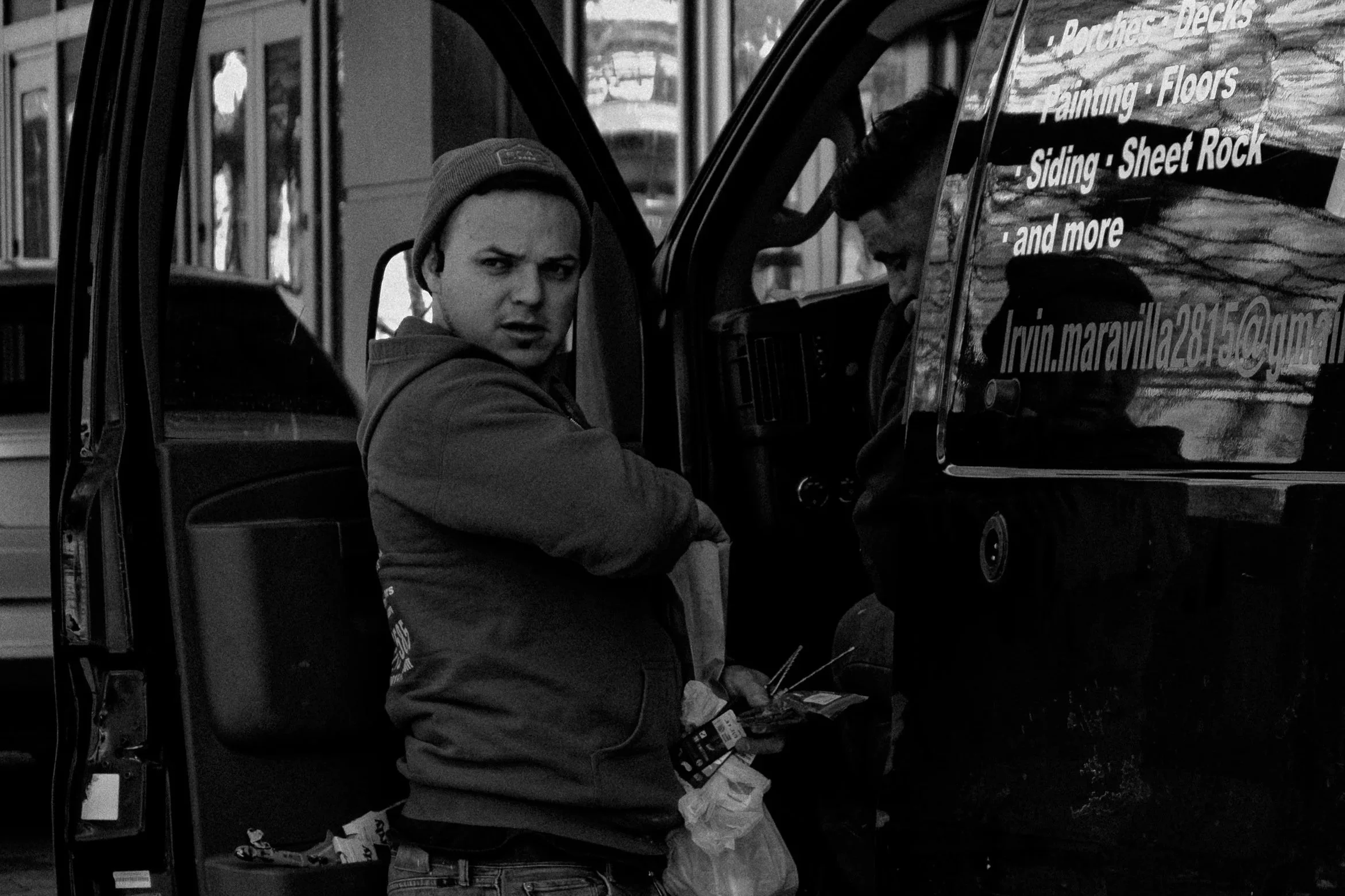 Man with a beanie and hoodie looking at the camera standing next to another person inside a vehicle, with business signage on the vehicle window.