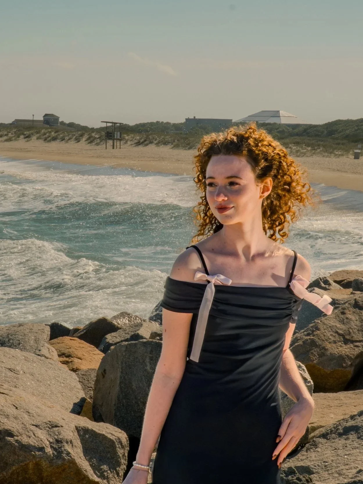 A woman with curly red hair in a black dress with pink ribbons on the shoulders standing on rocks at the beach, with ocean waves and sand in the background.