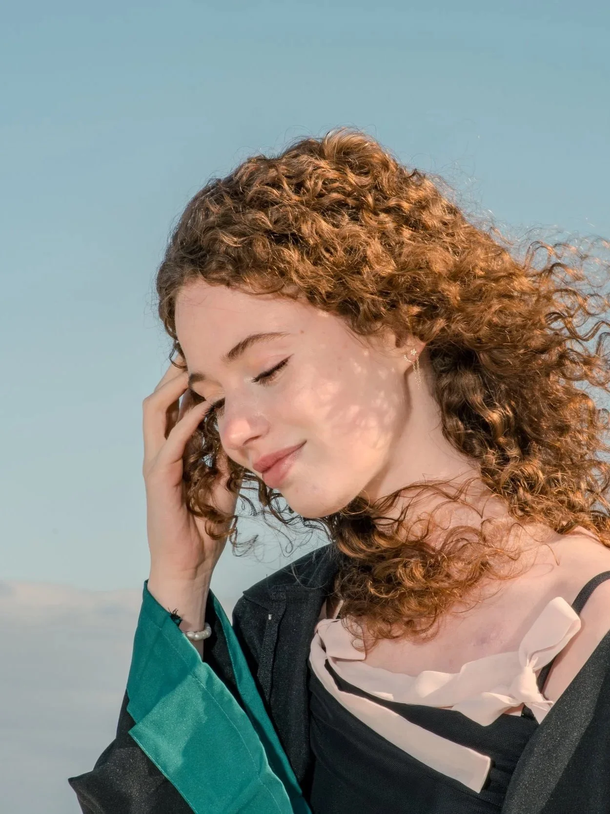 A woman with curly red hair, eyes closed, smiling softly, her hand resting on her temple, outdoors with a clear blue sky.