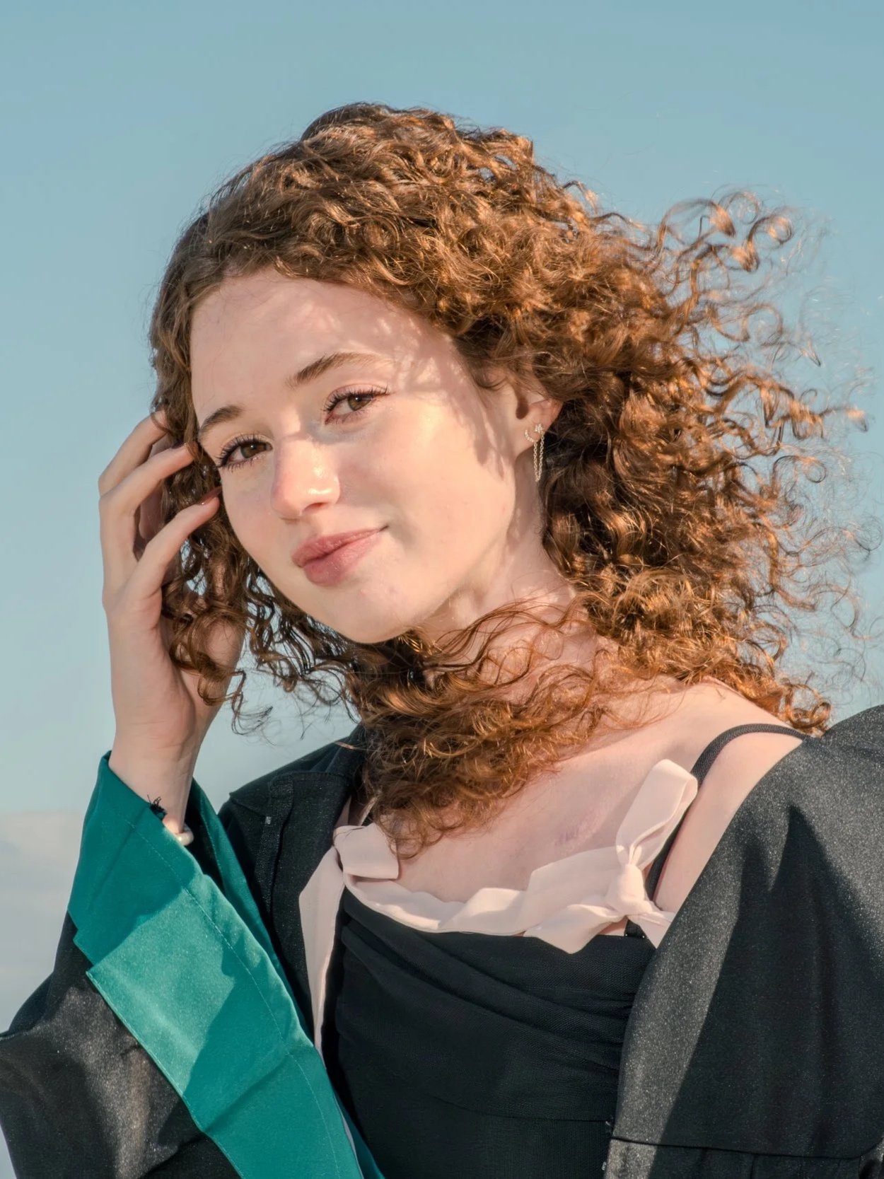 A young woman with curly red hair smiling outdoors against a clear blue sky, wearing a black outfit with a beige bow detail, a teal sleeve, and a small earring.