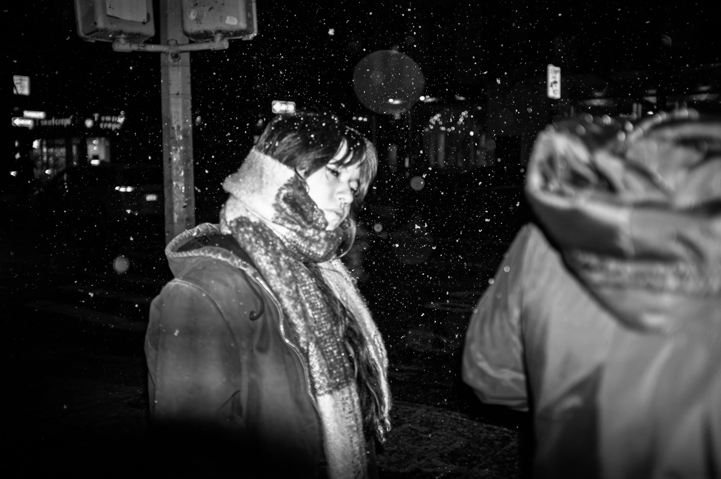 A woman in a coat and scarf standing in the snow at night, looking down, with snow falling around her. Part of another person is visible on the right.