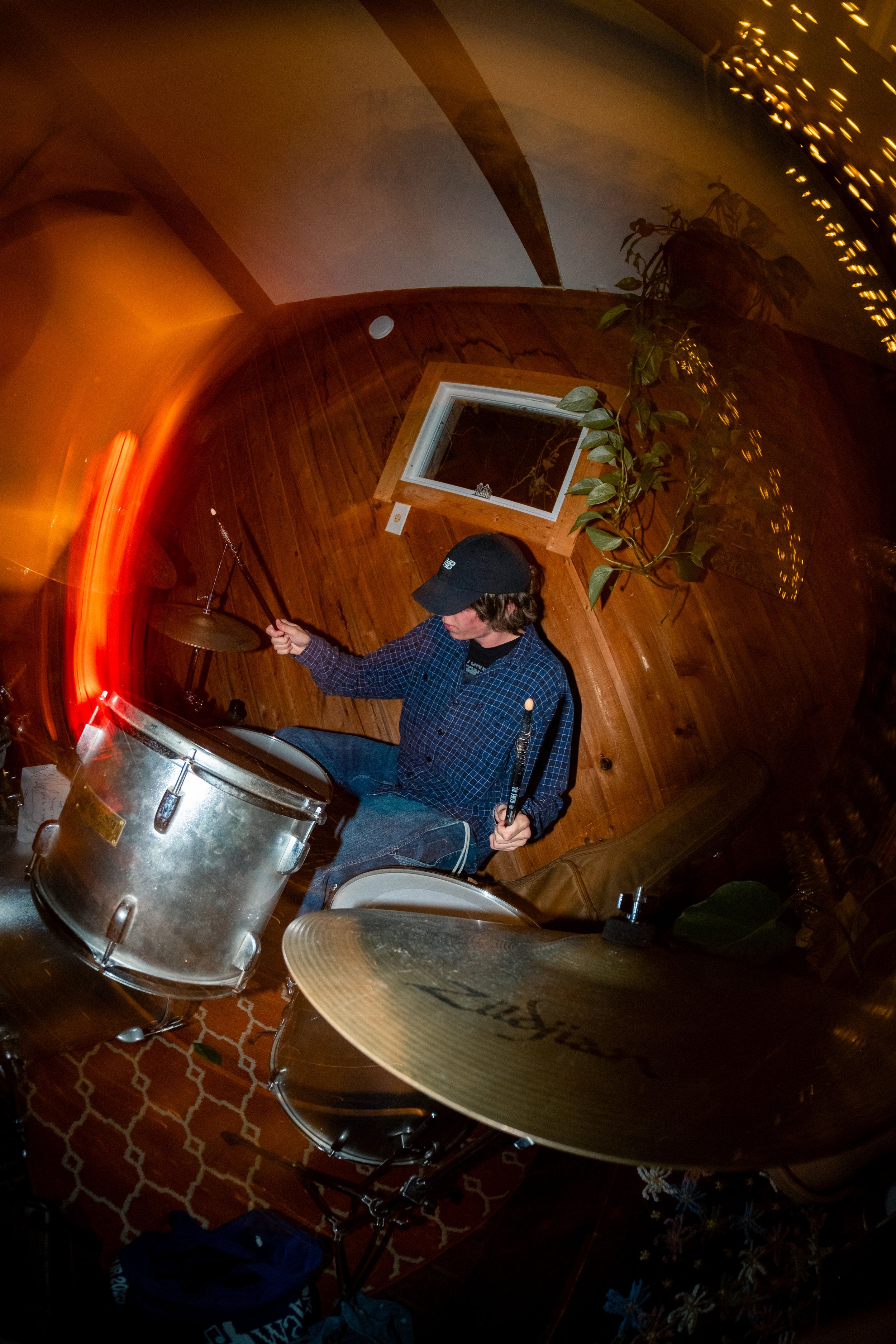 A person wearing a black cap and plaid shirt playing a drum set in a cozy wooden room decorated with string lights, a window, and plants.