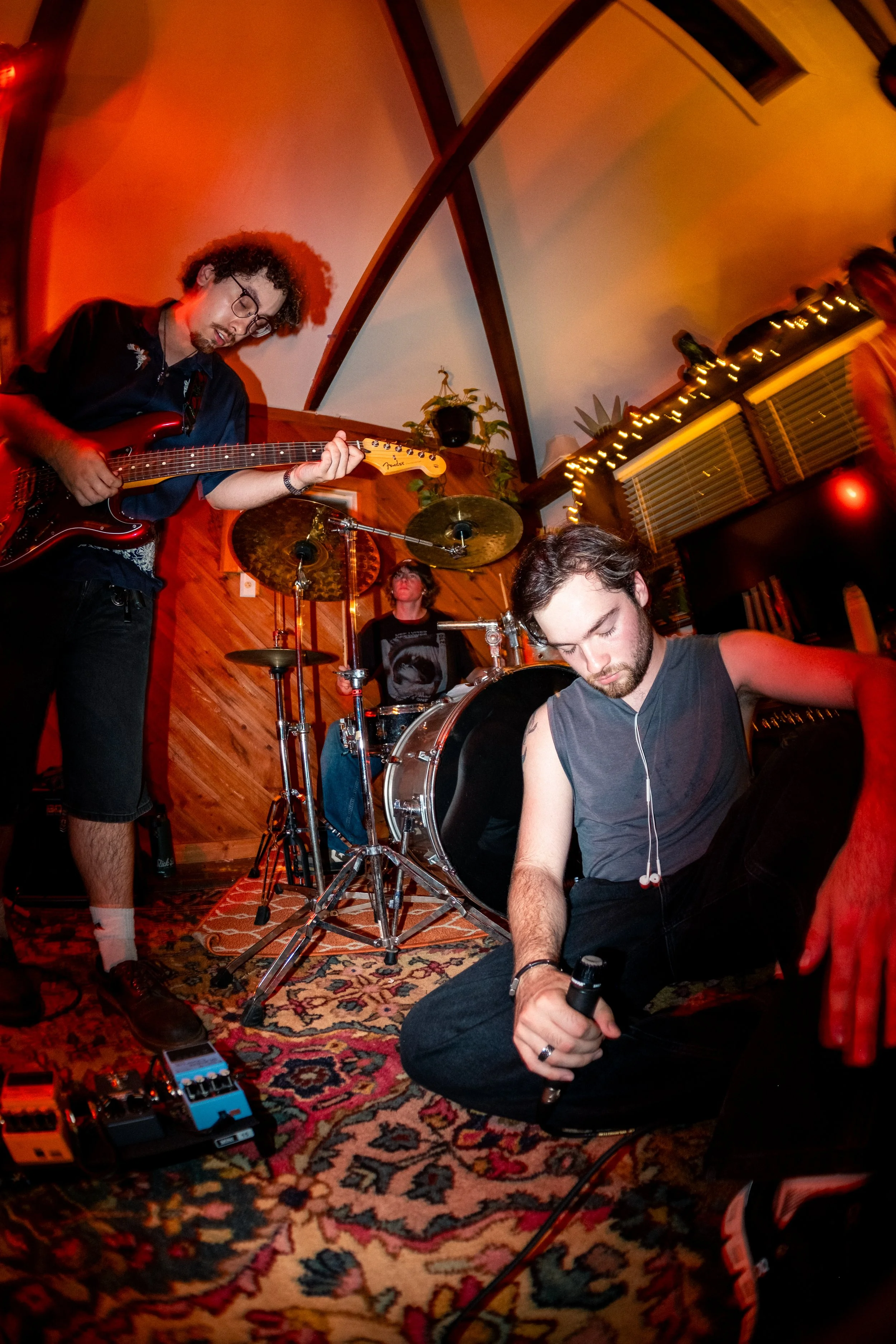 A group of musicians performing in a cozy, wood-paneled room with warm lighting and holiday decorations. The scene includes a guitarist, a drummer, and a singer with a microphone, surrounded by musical equipment and a detailed rug.
