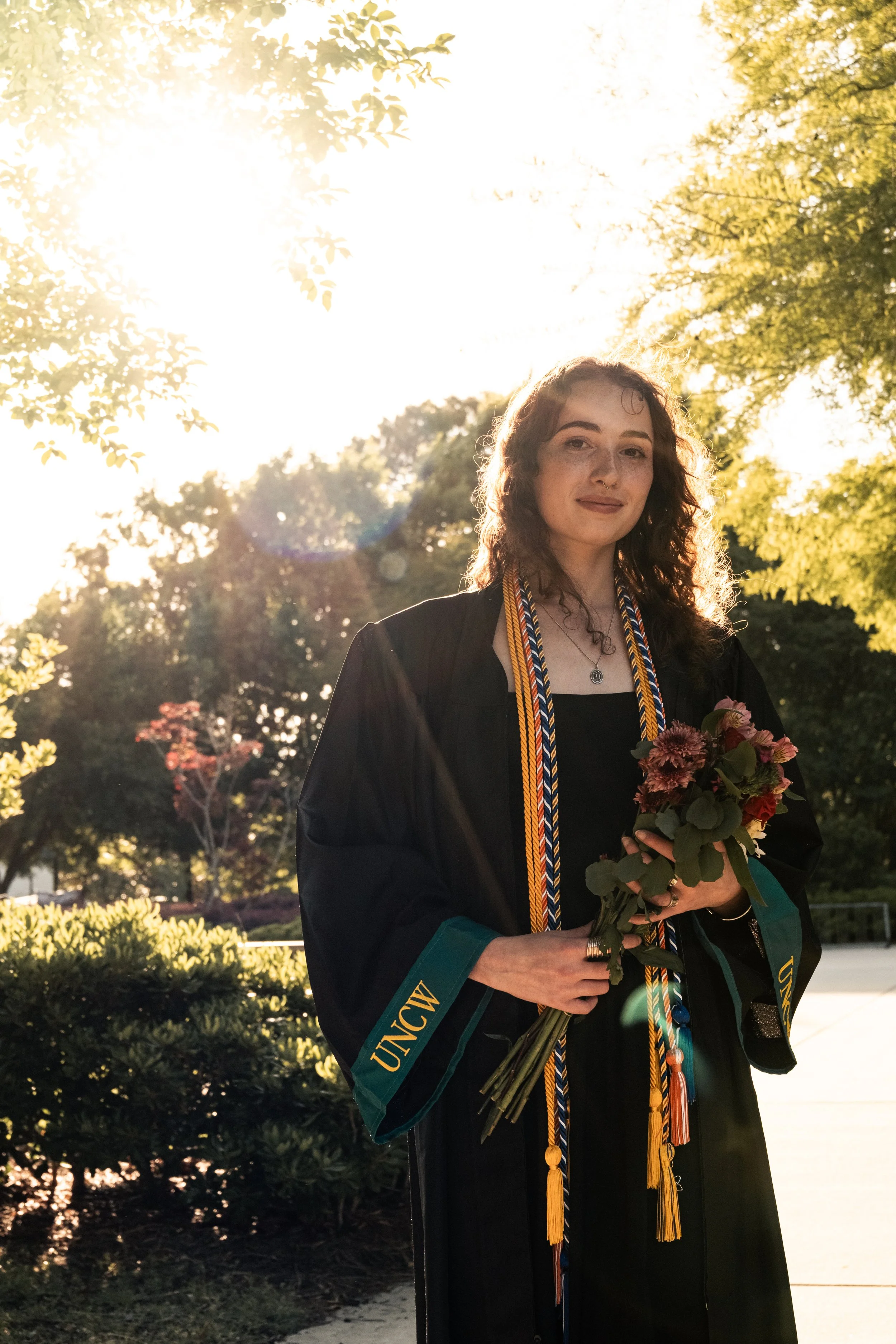 A woman in graduation attire holding a bouquet of flowers outdoor with sunlight behind her. She is wearing a black gown with green accents, a stole, and multiple honor cords. Trees and bushes are in the background.