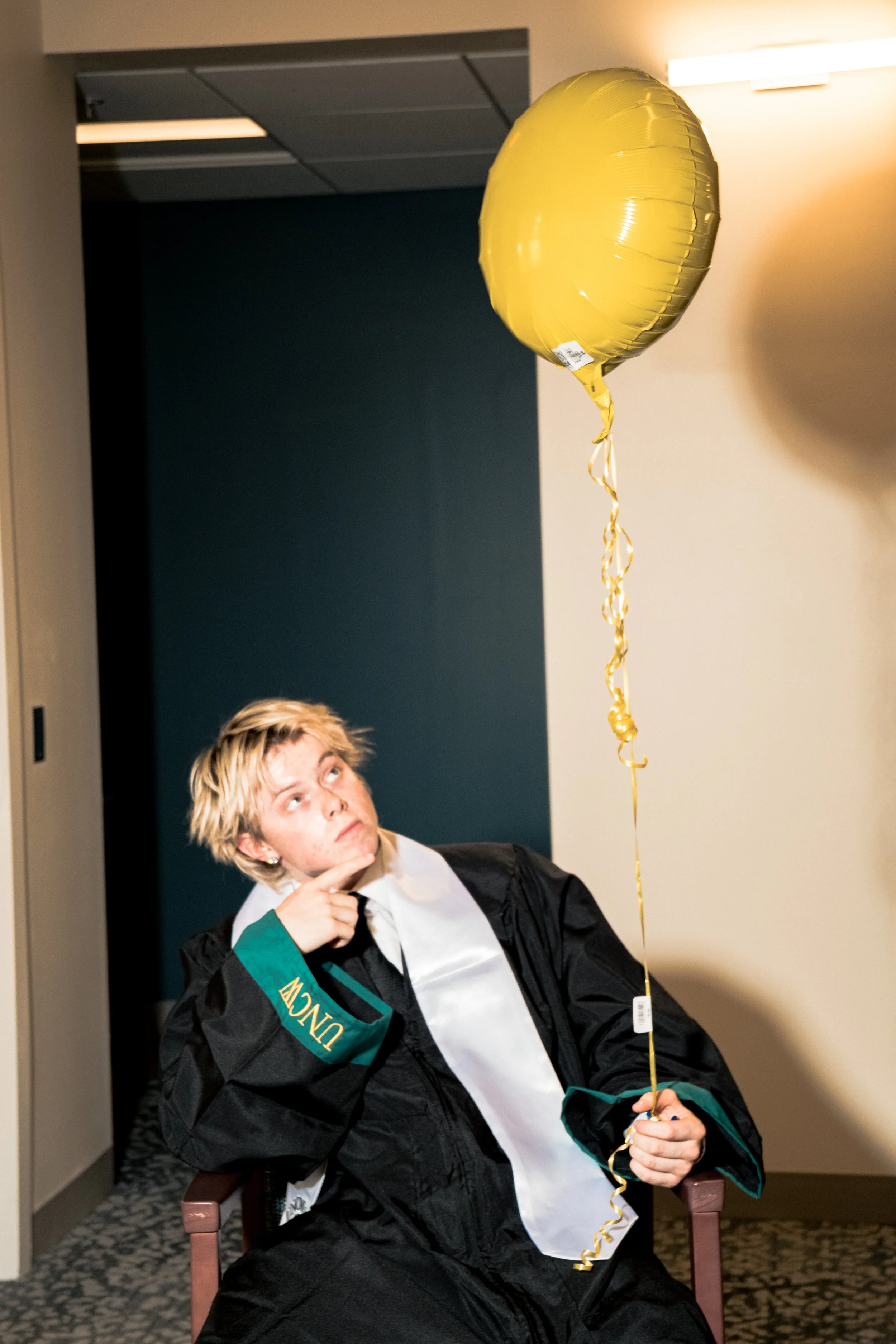 Person with blond hair in academic gown sitting in a chair, holding a yellow balloon, and thinking or posing for the photo indoors.