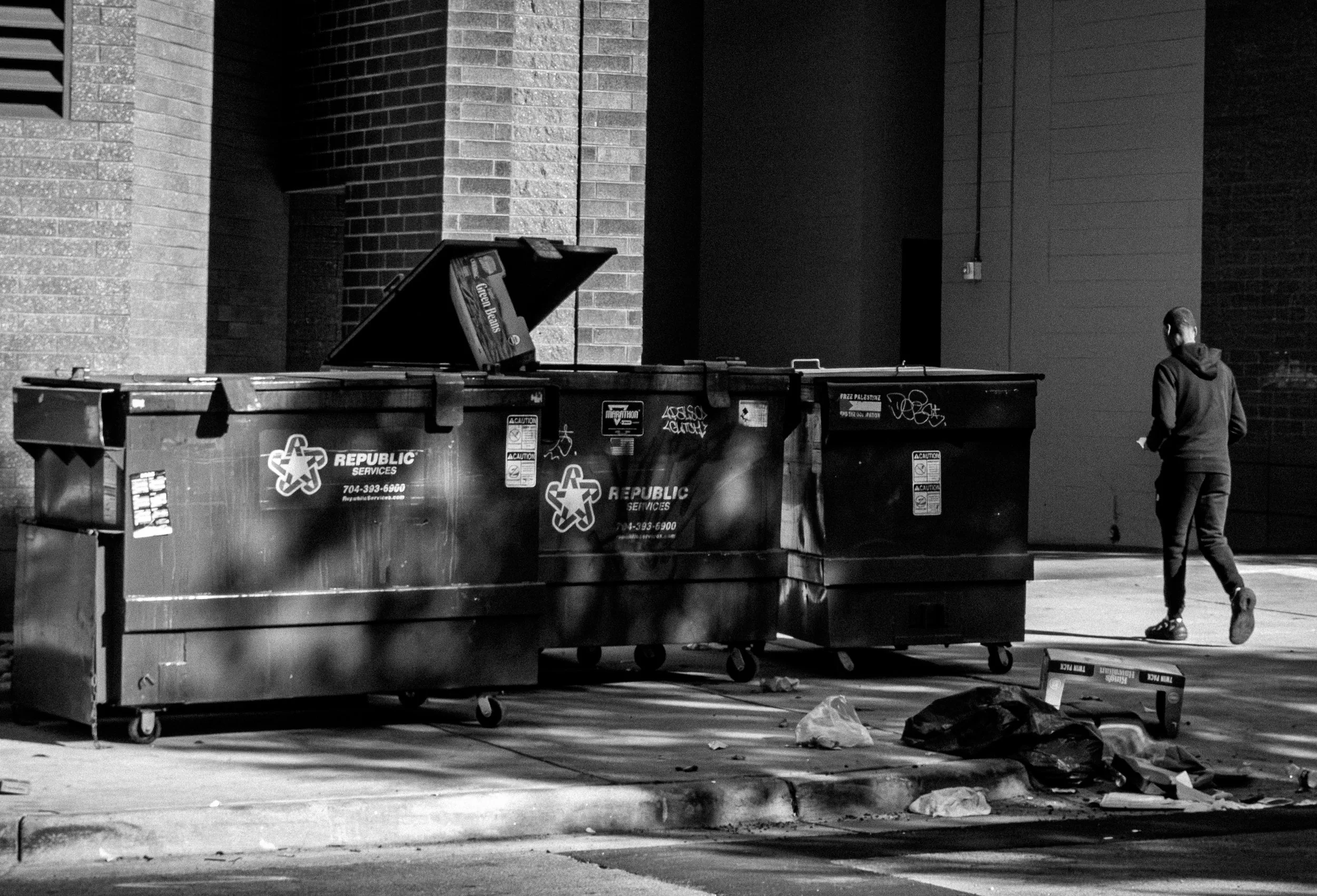 Nighttime scene of three dumpsters on a city sidewalk with a person walking past on the right. The dumpsters are marked with 'REPUBLIC SERVICES' logos and some graffiti, and trash is scattered on the ground nearby.