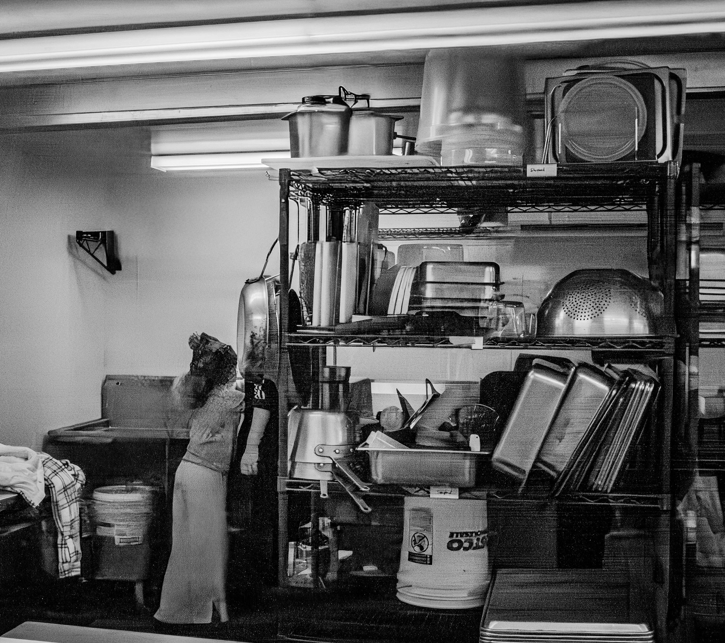 Storage room with kitchen equipment such as trays, pots, lids, and a large bucket, with a woman standing near a table covered with clothes.