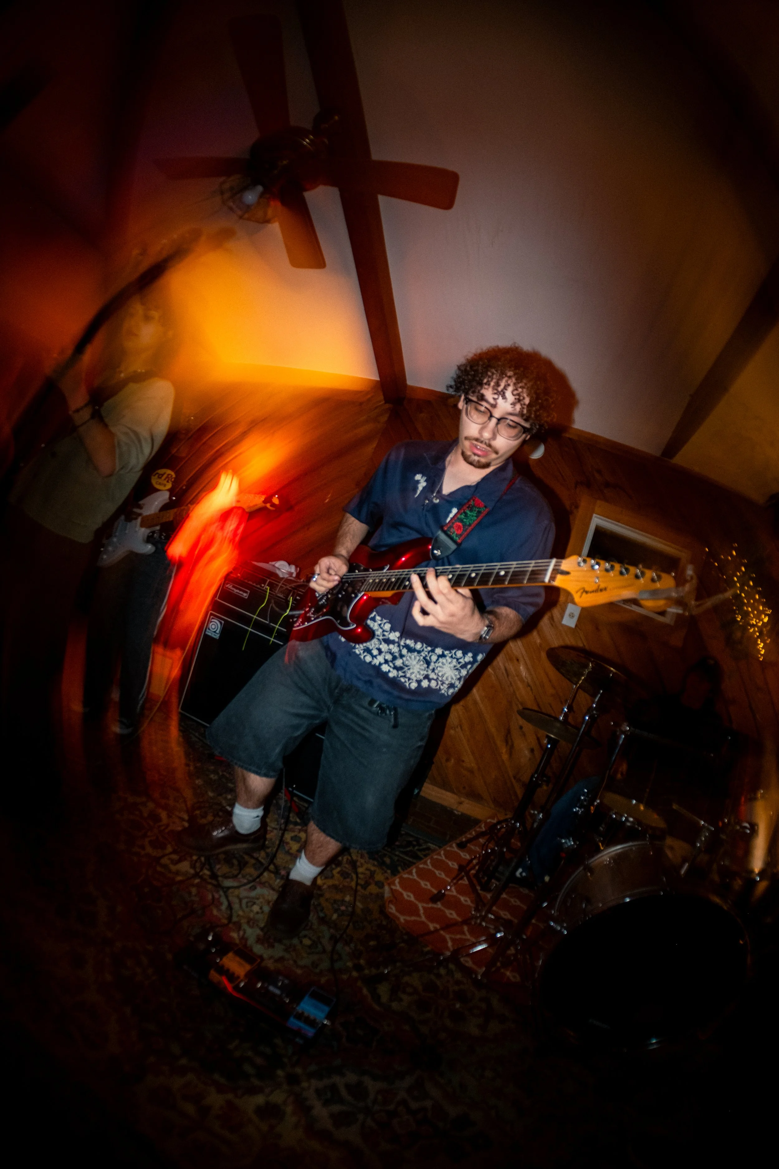 A young man with curly hair, glasses, and a goatee playing an electric guitar in a wooden-paneled room. He is wearing a short-sleeved shirt, shorts, and white socks with brown shoes. There is a drum set and music equipment nearby, and a person with a