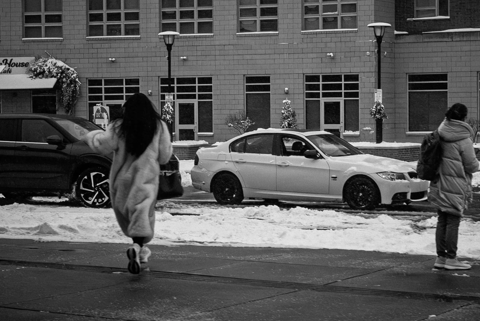 A black and white photo of a snowy city street scene with two women walking on the sidewalk, with parked cars and a building facade in the background.