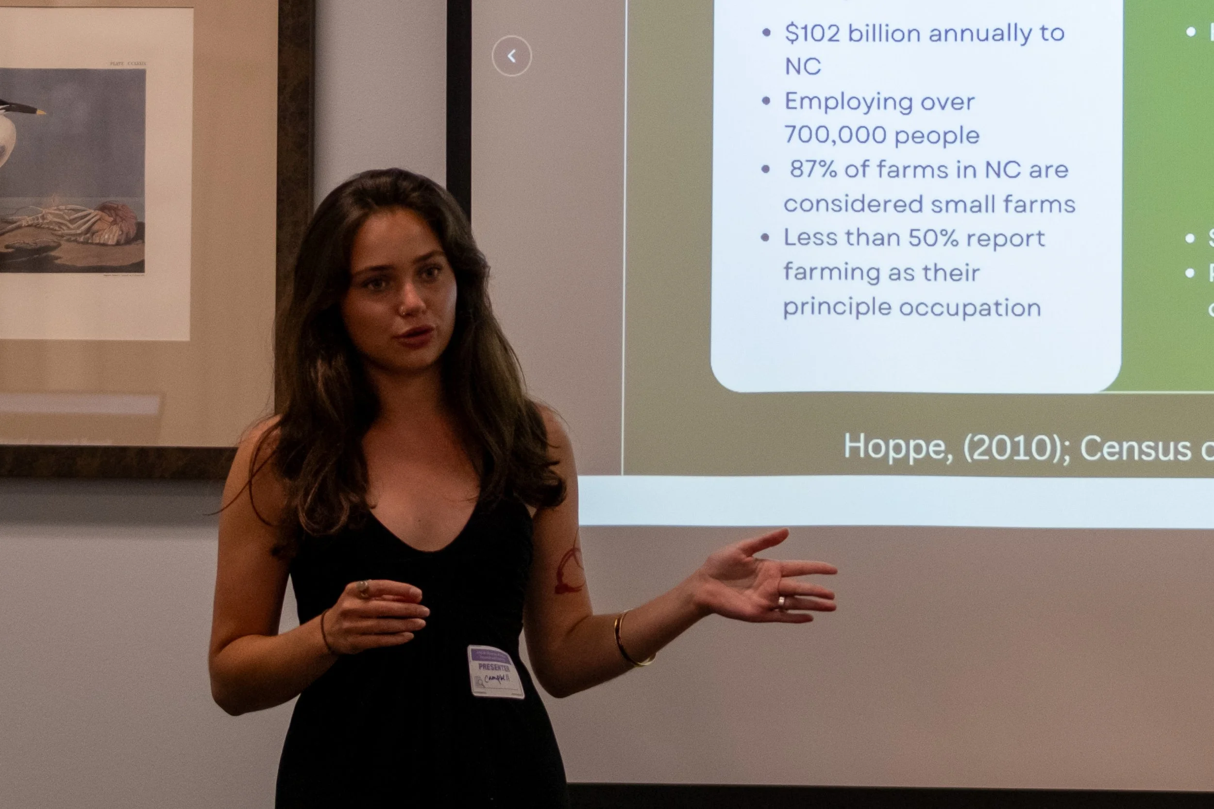 A woman with long brown hair, wearing a black sleeveless top and a name tag, presenting in front of a projection screen displaying text about farm employment and economics. She gestures with her right hand, explaining the slide.