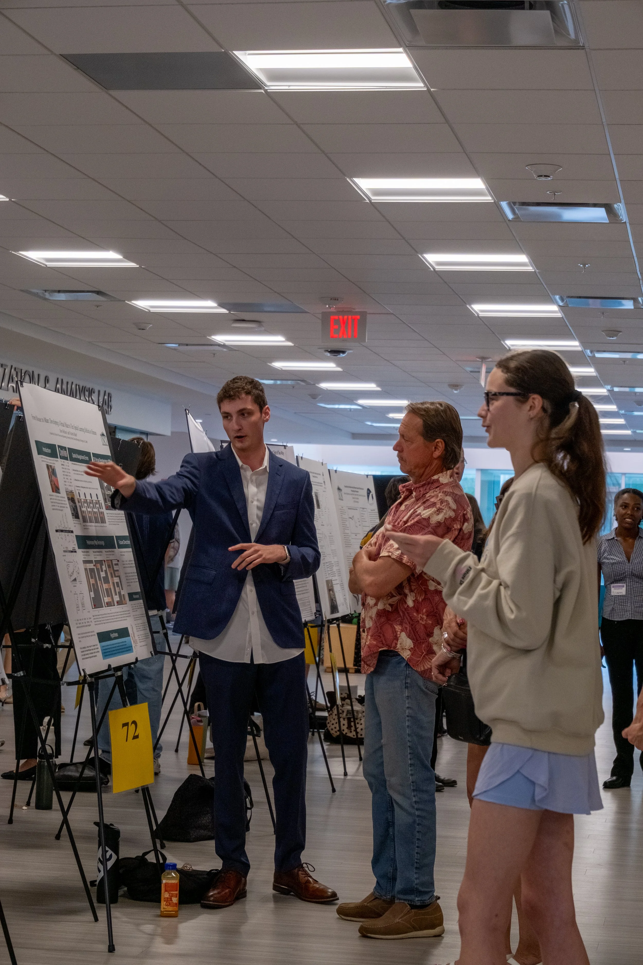 Three people are attending a scientific poster presentation in an indoor setting with a ceiling of square tiles and several rectangular light fixtures. The man on the left, wearing a dark suit and white shirt, is explaining the poster to the two wome