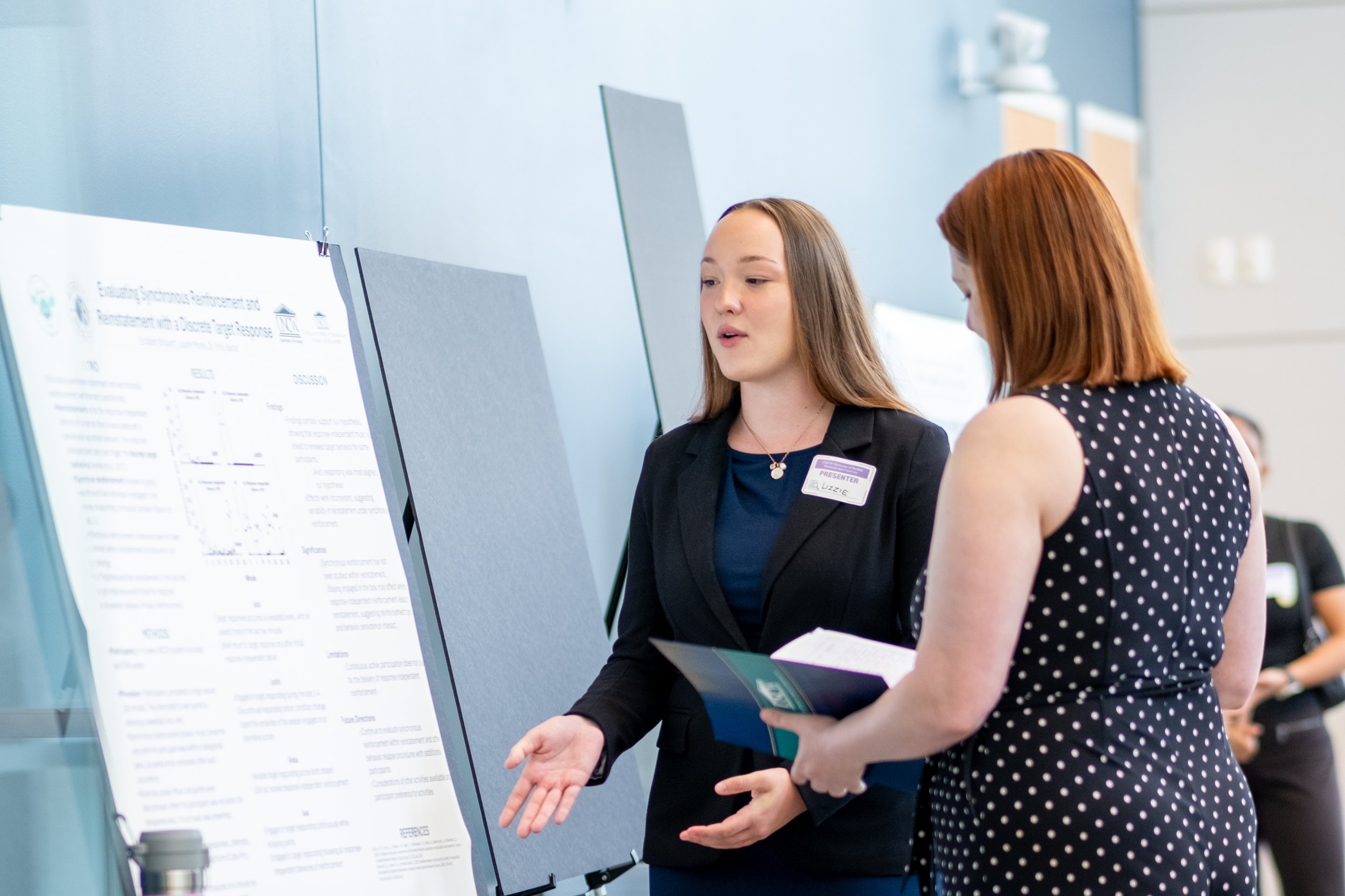 Two women, one with long brown hair and another with short red hair, are engaged in conversation at a scientific or academic conference, standing in front of a presentation board with text and diagrams.