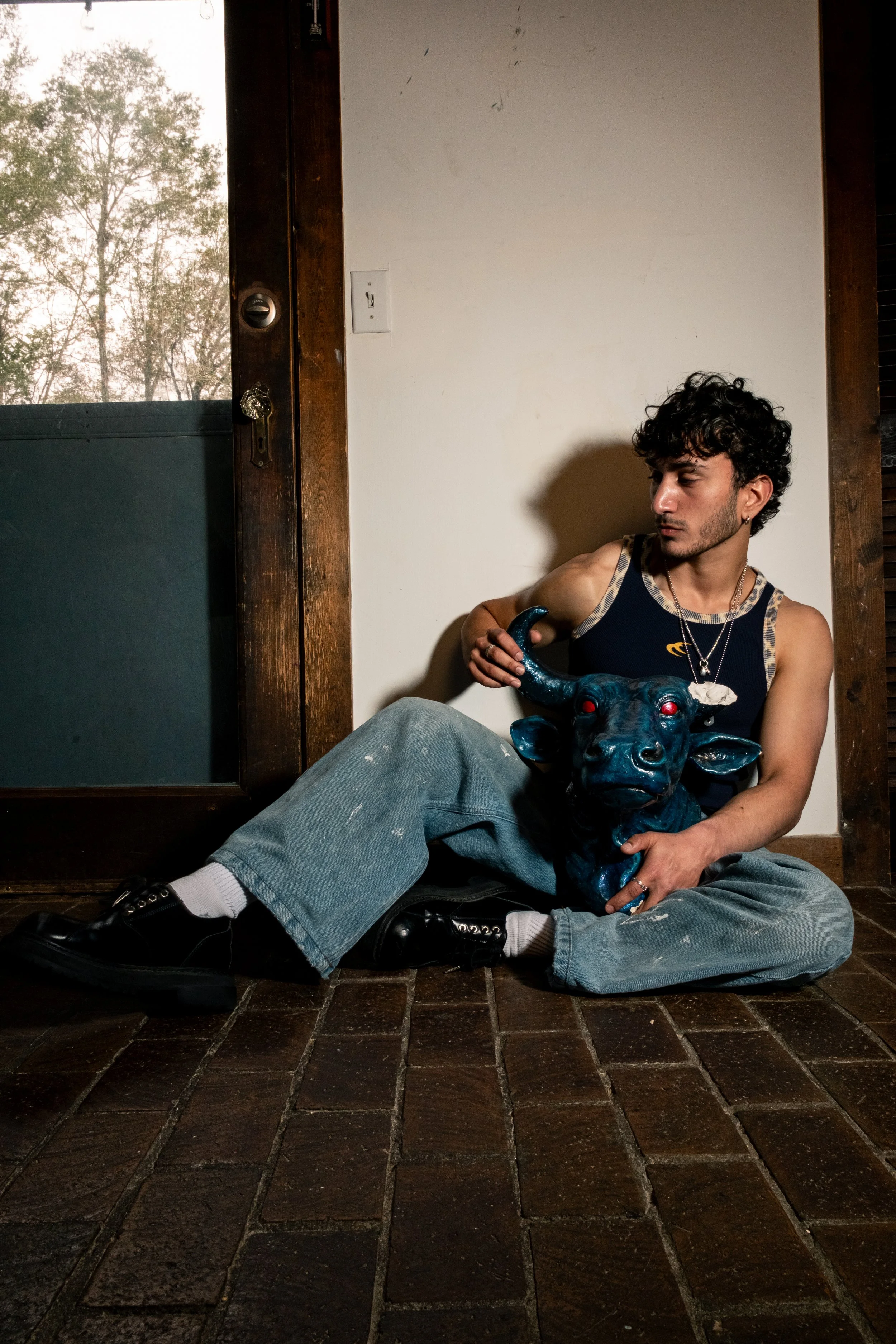 A young man with curly dark hair, wearing a sleeveless shirt, sitting on a tiled floor against a white wall, holding a shiny blue statue of a bull with red eyes. There is a window with a view of trees behind him.