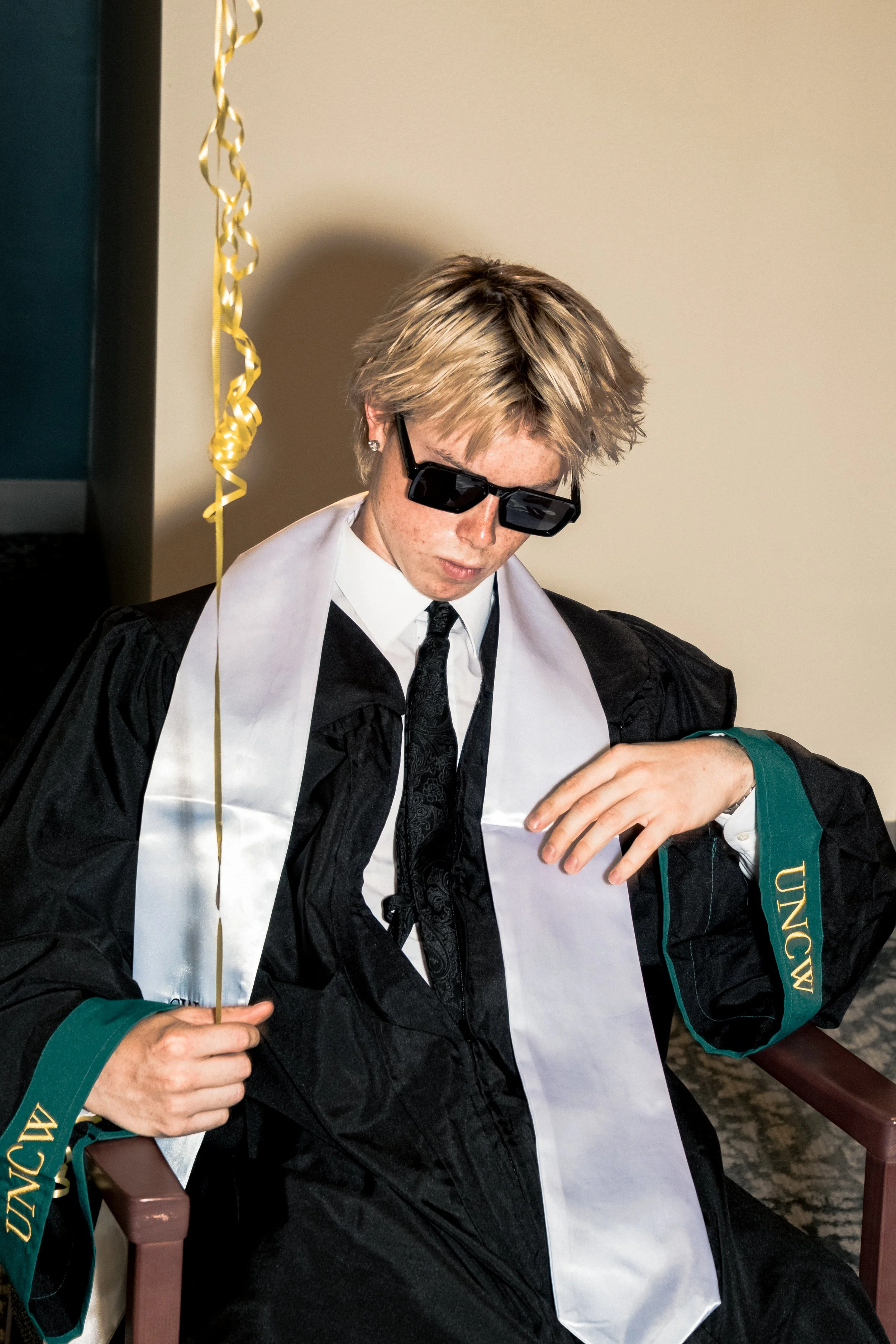 A young man in academic regalia, including a black gown, white stole, and a green sleeve with gold embroidery, is sitting and looking down. He is wearing black sunglasses and has blonde, messy hair. He appears to be at a graduation ceremony, holding 