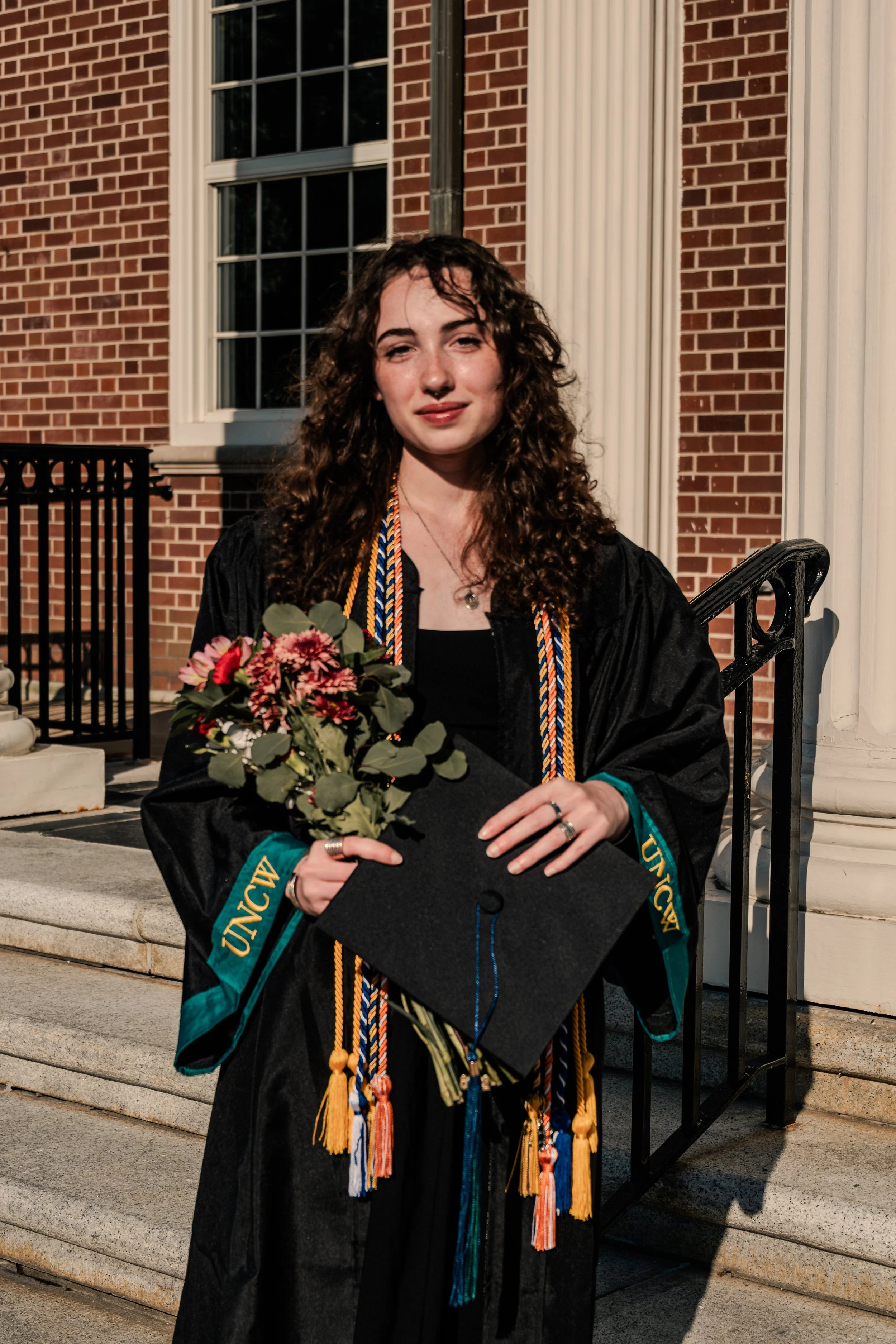 A young woman with curly hair in graduation cap and gown holding a bouquet of flowers outside a brick building.