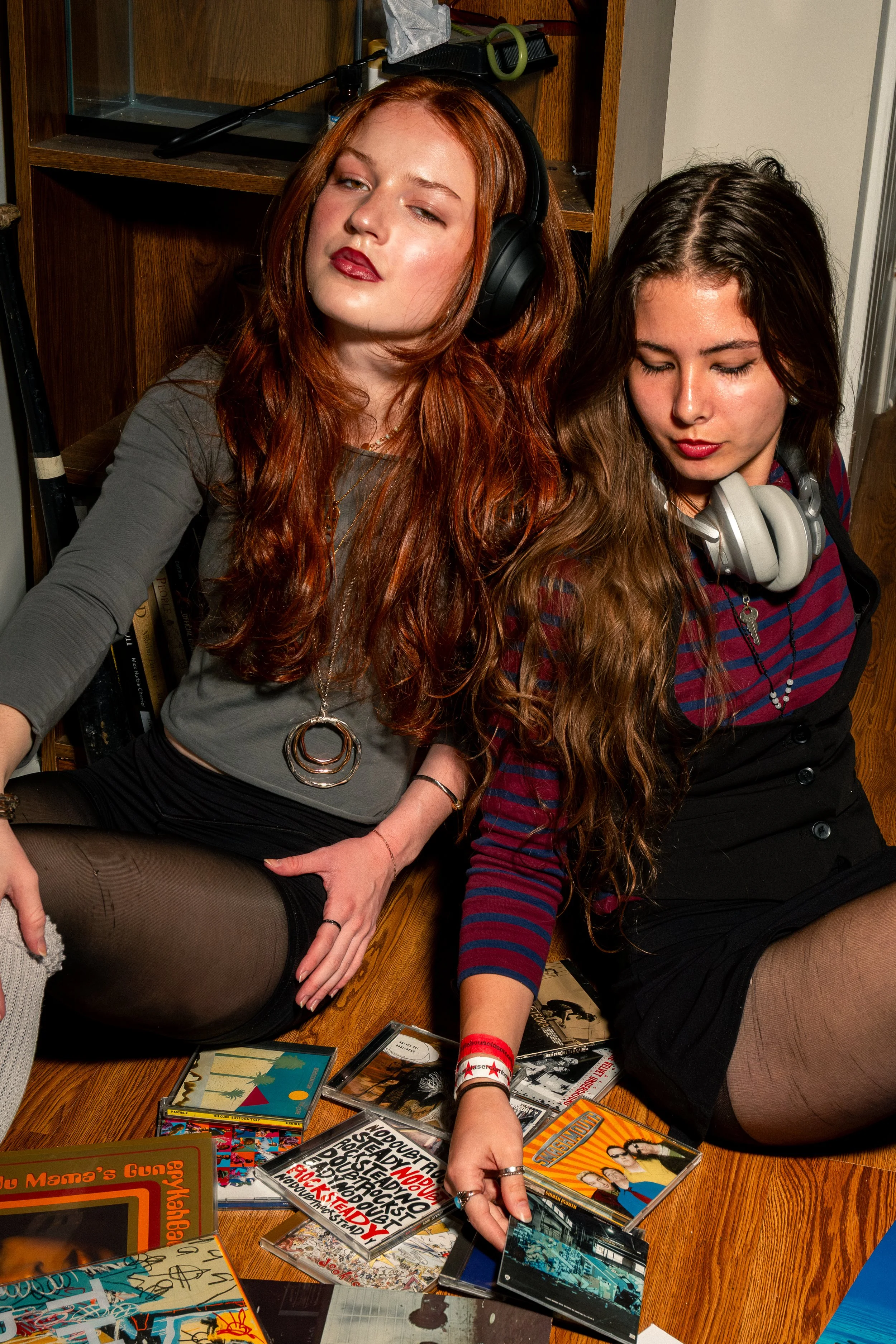 Two young women with long hair, one with red hair and the other with brown hair, are sitting on the floor with a collection of CDs and DVDs spread out in front of them. The woman on the left is wearing a gray top, a large necklace, and black stocking