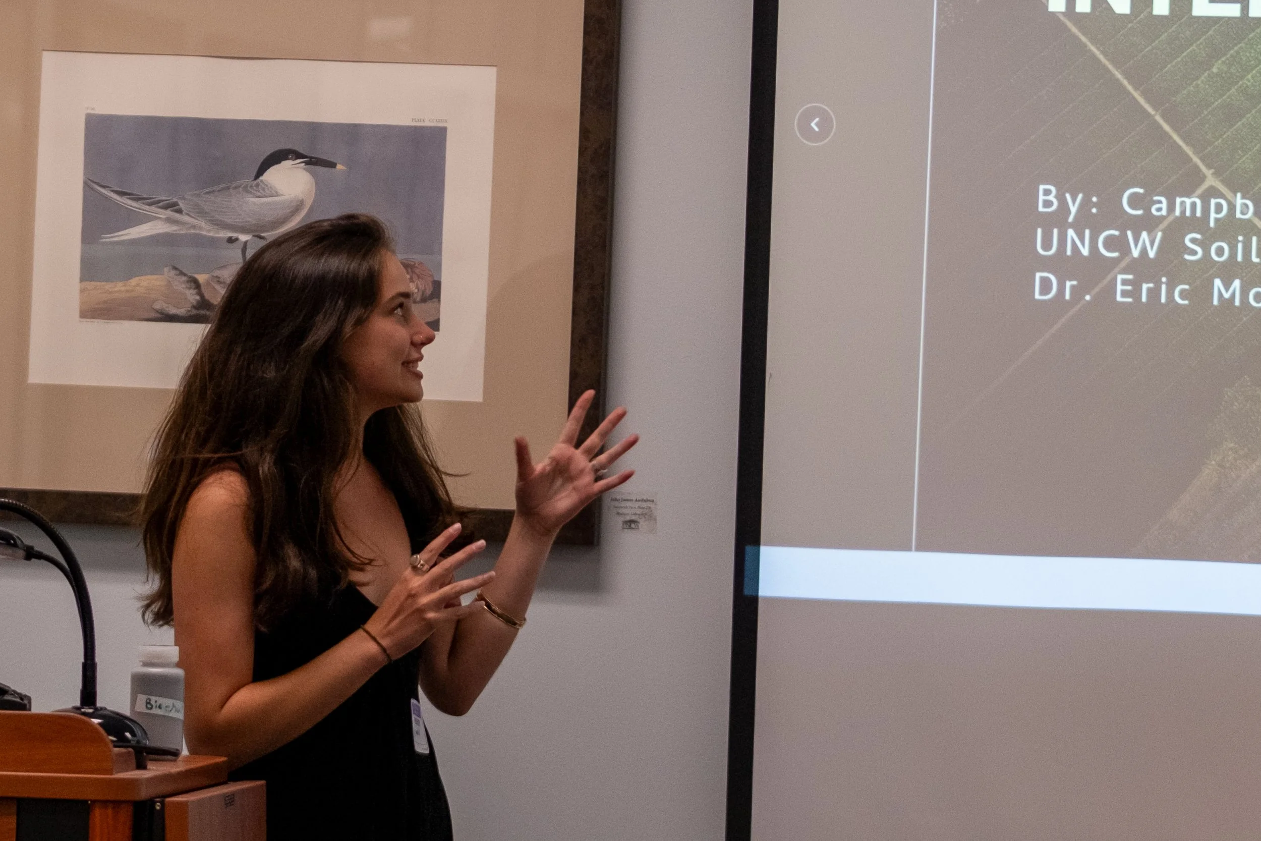 A woman with long brown hair wearing a black top and a ring on her finger is gesturing with her hands while standing in front of a presentation screen. The screen displays a presentation titled 'By: Камб' with additional text that is partially visibl