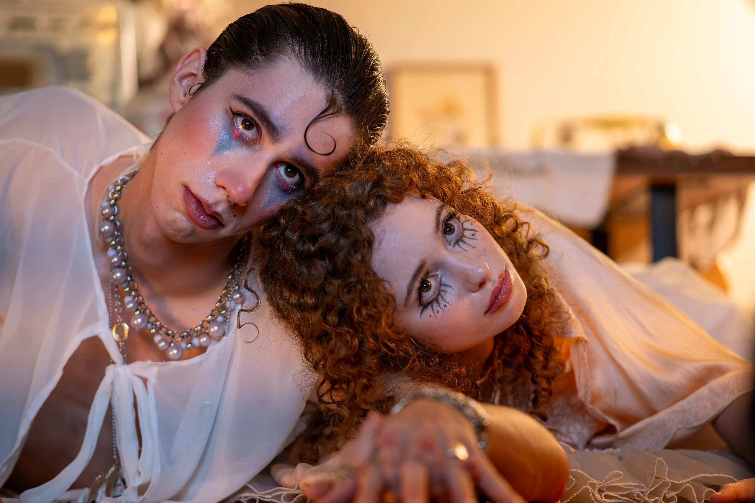 Two young women with dramatic makeup and curly hair lying close together on a bed, staring into the camera. One woman has black hair and wears a pearl necklace, the other has reddish-brown hair and animated eye makeup, with a background of a cozy, warmly lit room.