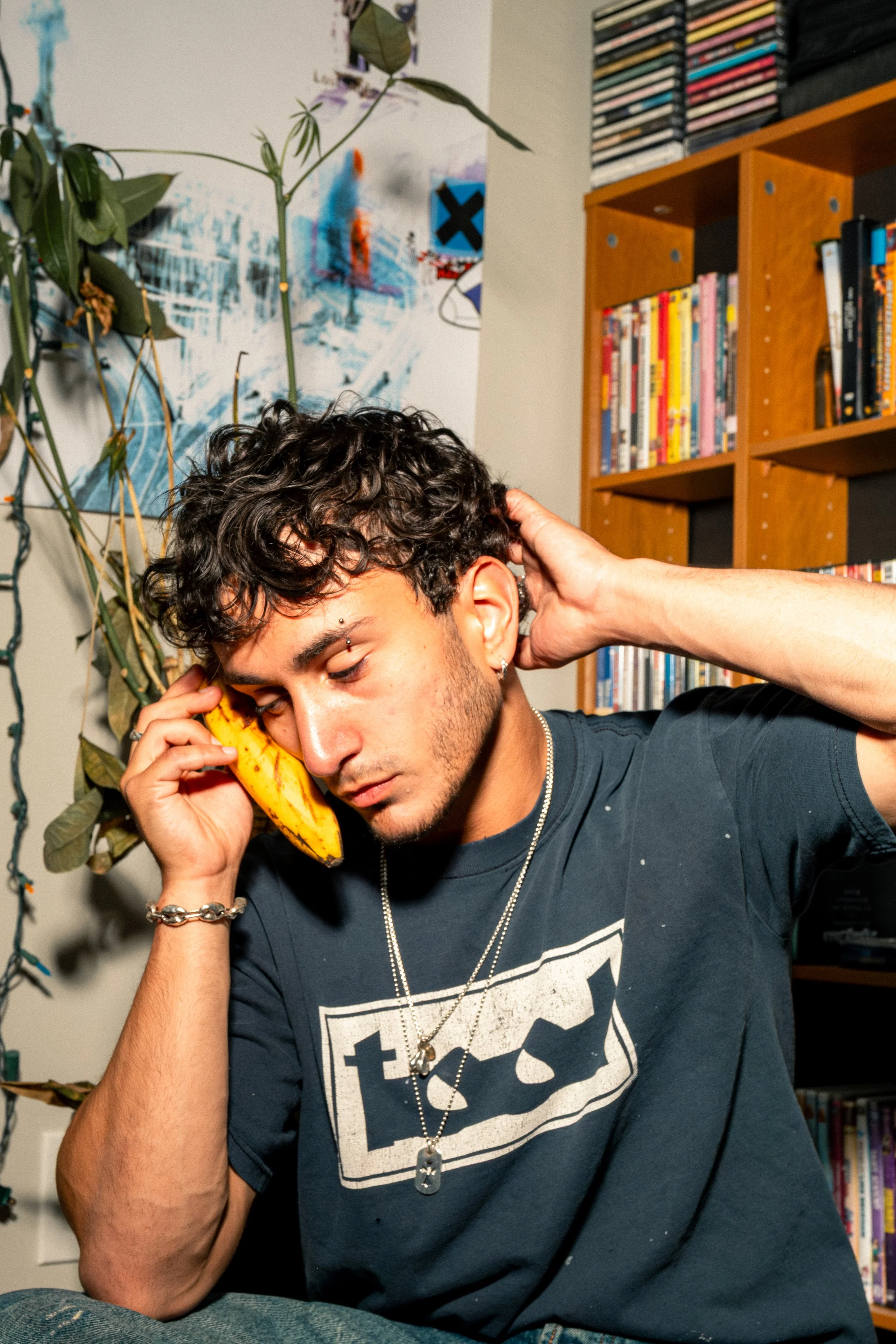 A young man with curly dark hair talking on a banana-shaped phone, wearing a dark graphic t-shirt with a septum piercing and layered necklaces, sitting in front of a bookshelf with colorful books, a poster on the wall, and a potted plant.