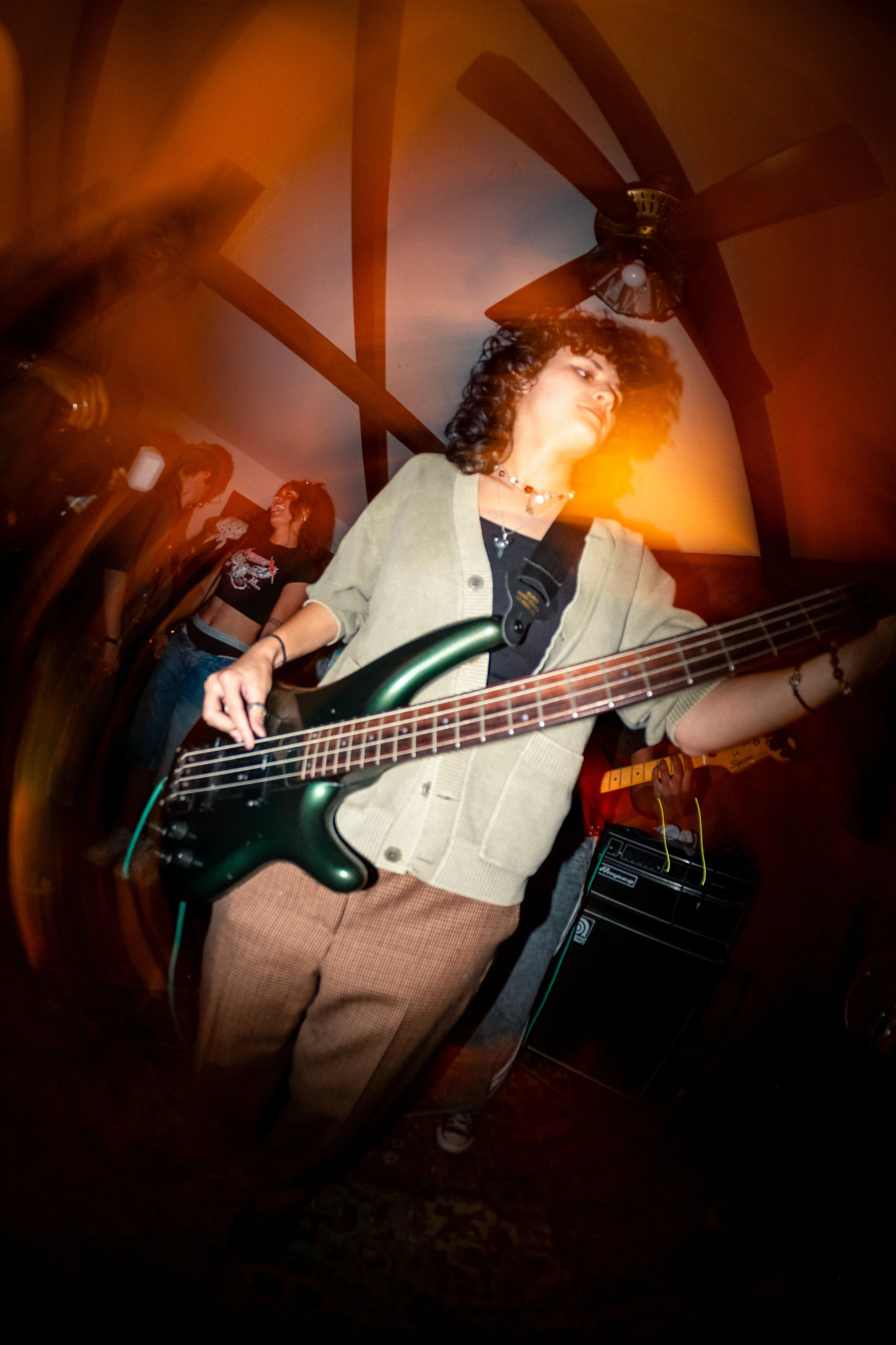 A woman with curly hair playing a green electric bass guitar at a music event, with a woman smiling and talking in the background, under warm lighting in an indoor setting.