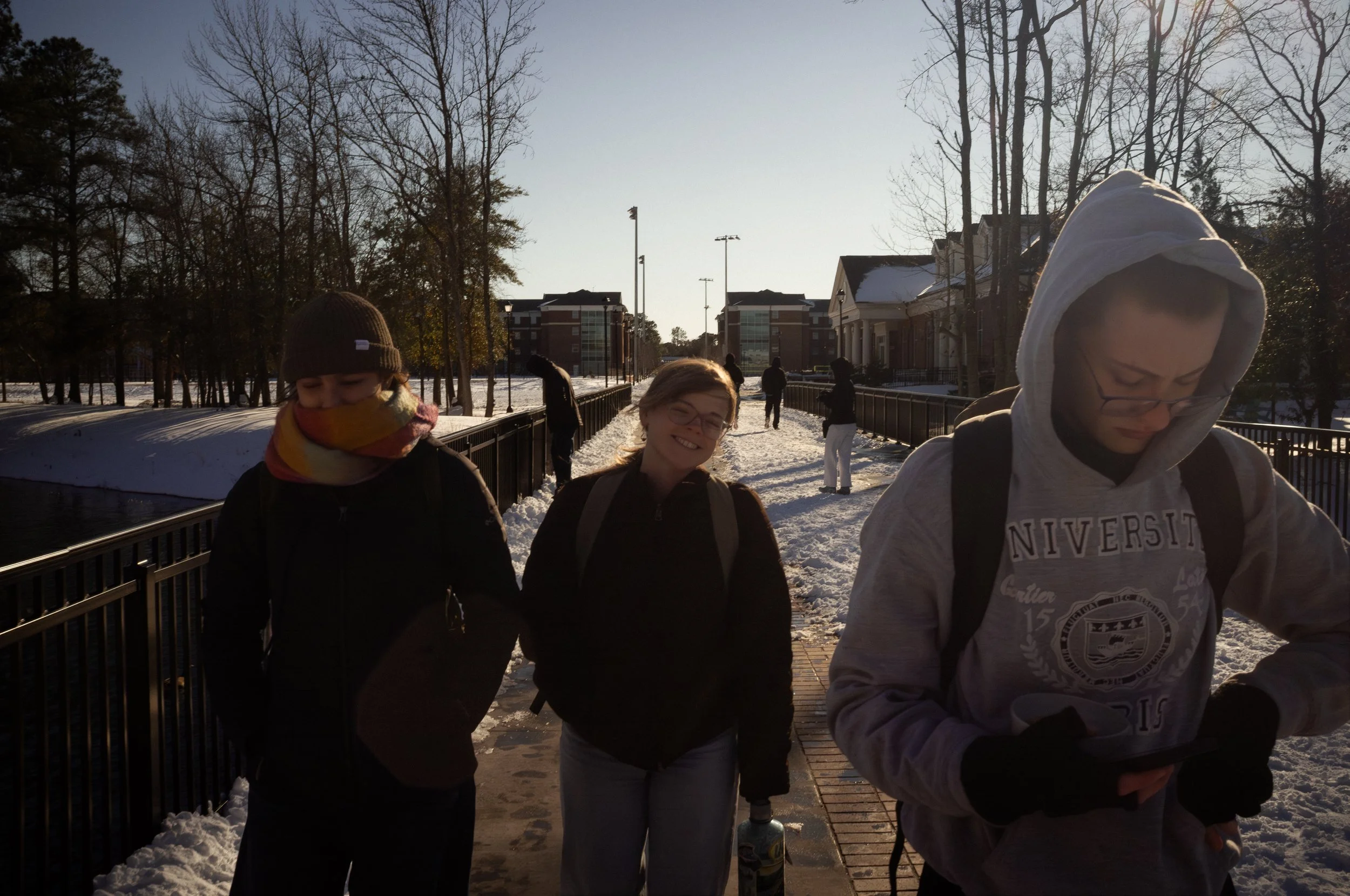 Group of four young people walking outdoors on a snowy pathway during the daytime, with leafless trees and buildings in the background.
