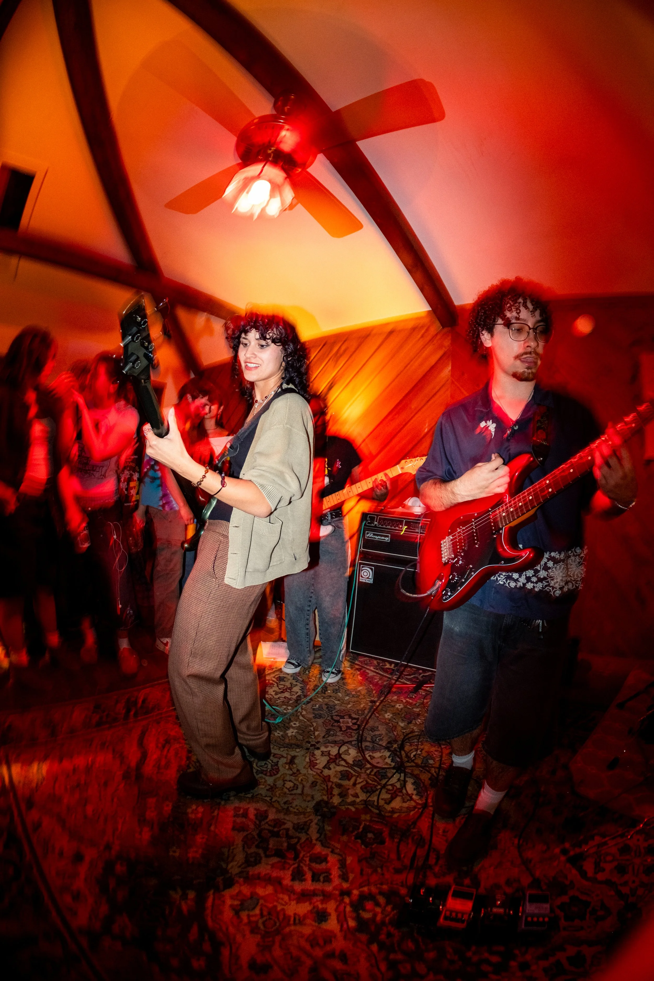 A group of musicians performing with guitars in a room with a wooden ceiling and a ceiling fan, and a group of people dancing or watching in the background.