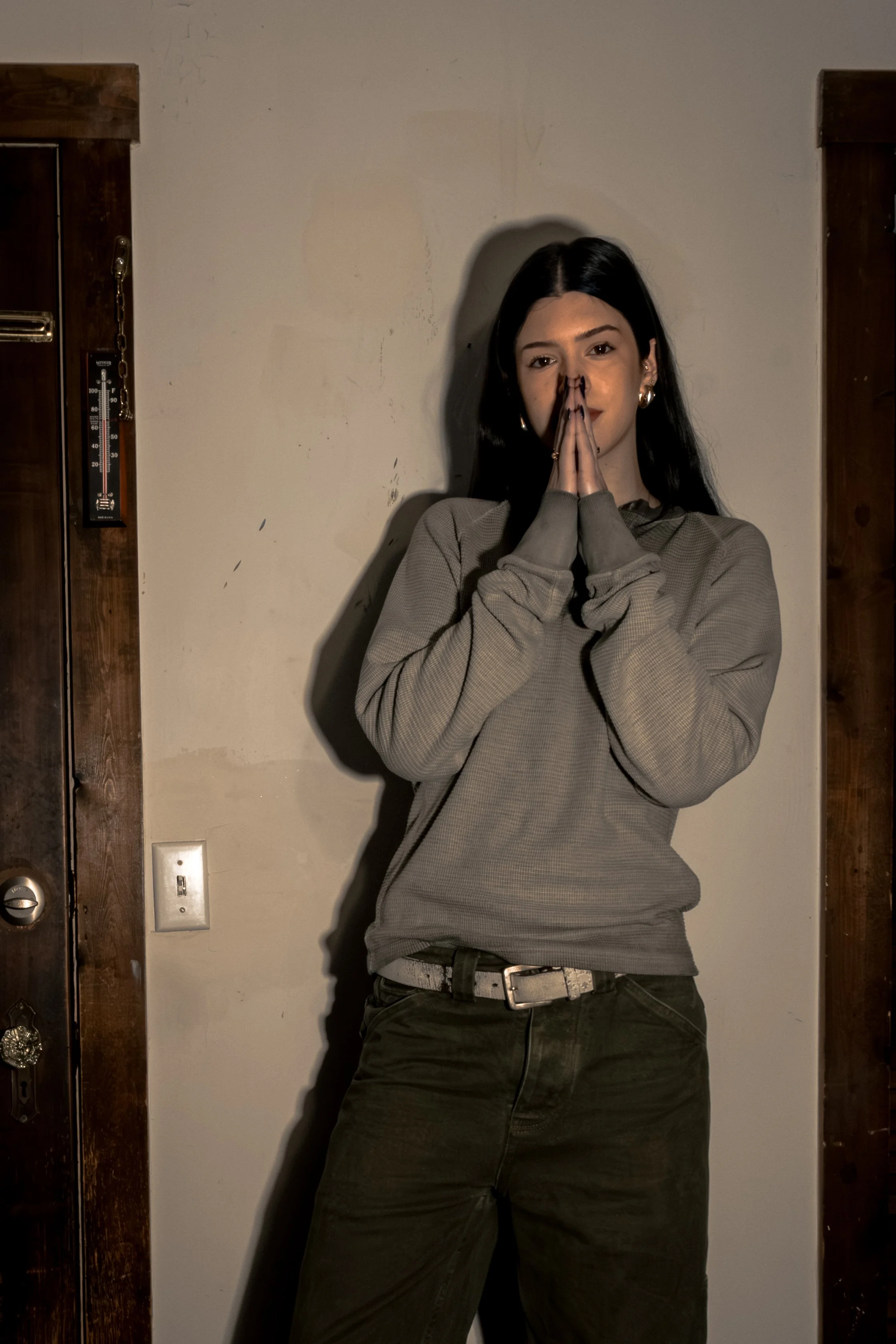 Young woman with long black hair and hoop earrings standing indoors with hands pressed together in prayer gesture, wearing a gray long sleeve shirt and black pants.